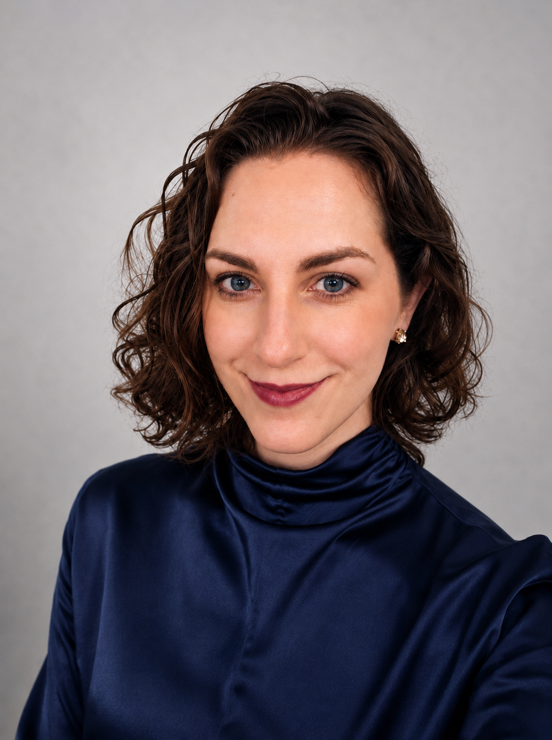 A young woman with curly brown hair and blue eyes taking a selfie against a light gray background. She is wearing a high-necked dark blue satin blouse and gold earrings, and is smiling softly.
