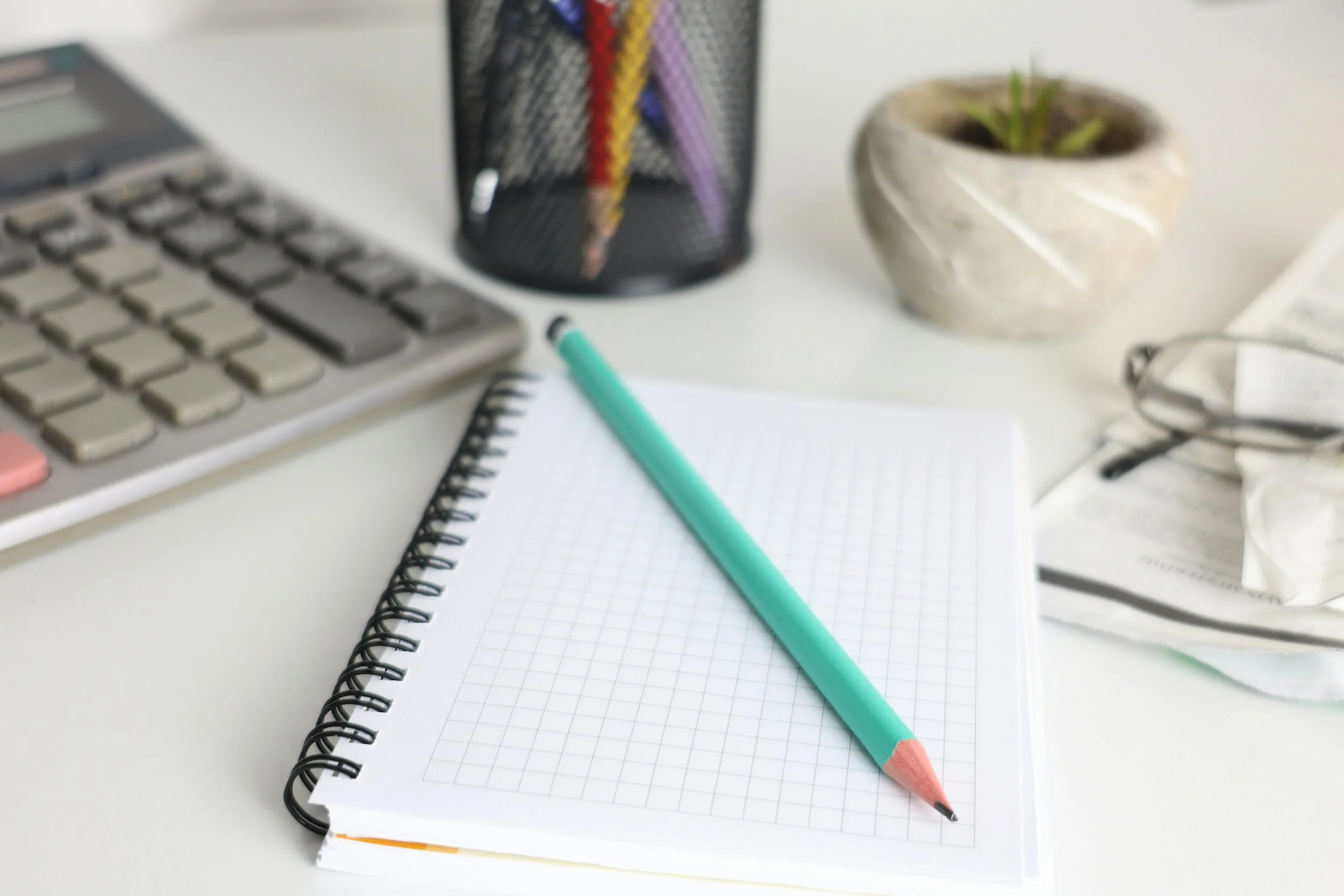 Close-up of a white desk with a spiral notebook with graph paper, a mint green pencil with a pink eraser, a calculator, a black pen holder with colorful pencils, a potted plant, a pair of glasses, and some scattered papers.