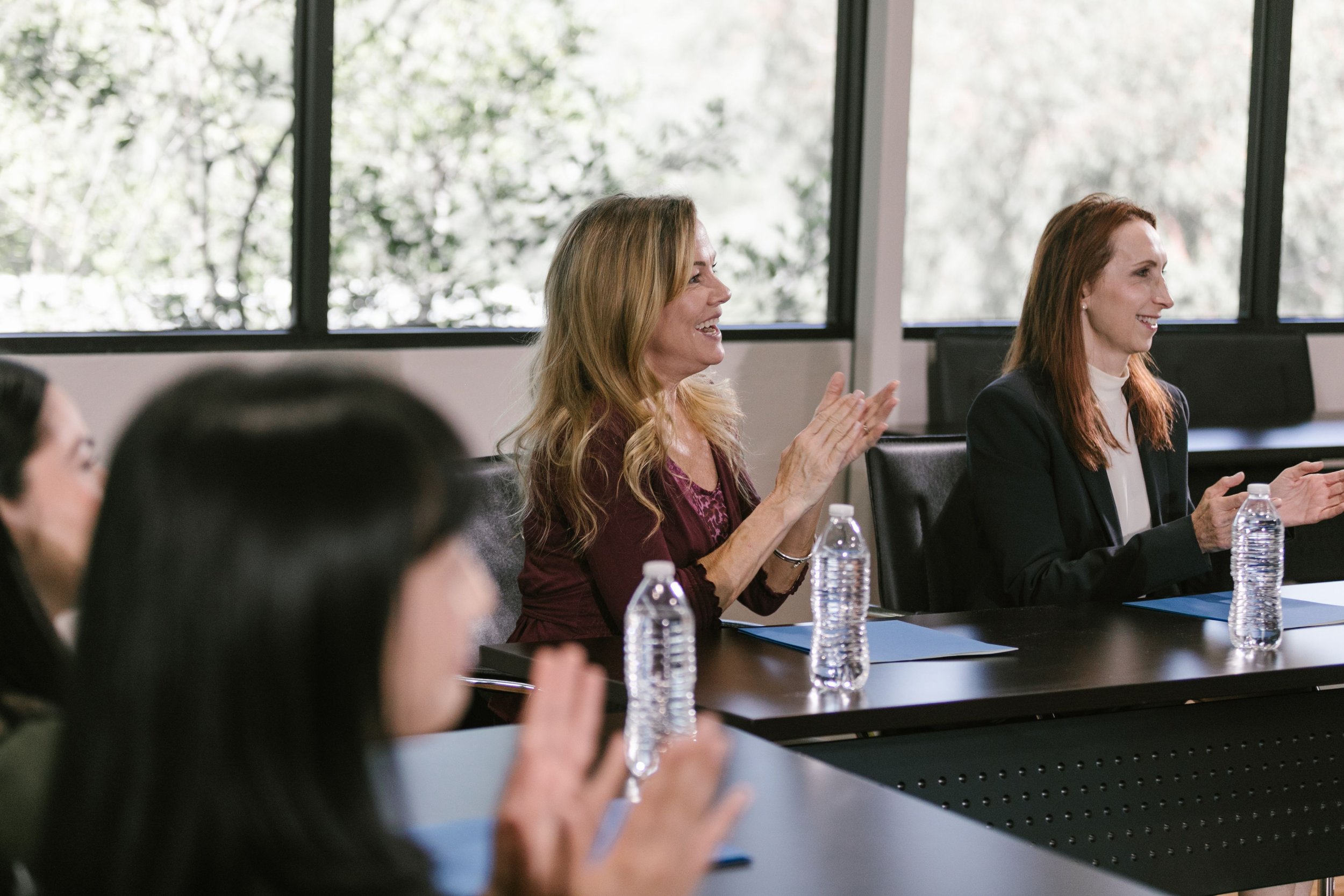 Women attending a meeting, sitting at a conference table, smiling, with water bottles in front of them, in a room with large windows showing trees outside.