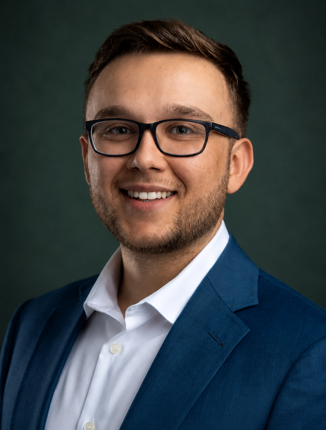Headshot of a young man with short brown hair, glasses, a beard, wearing a navy blue suit and white shirt, smiling at the camera.