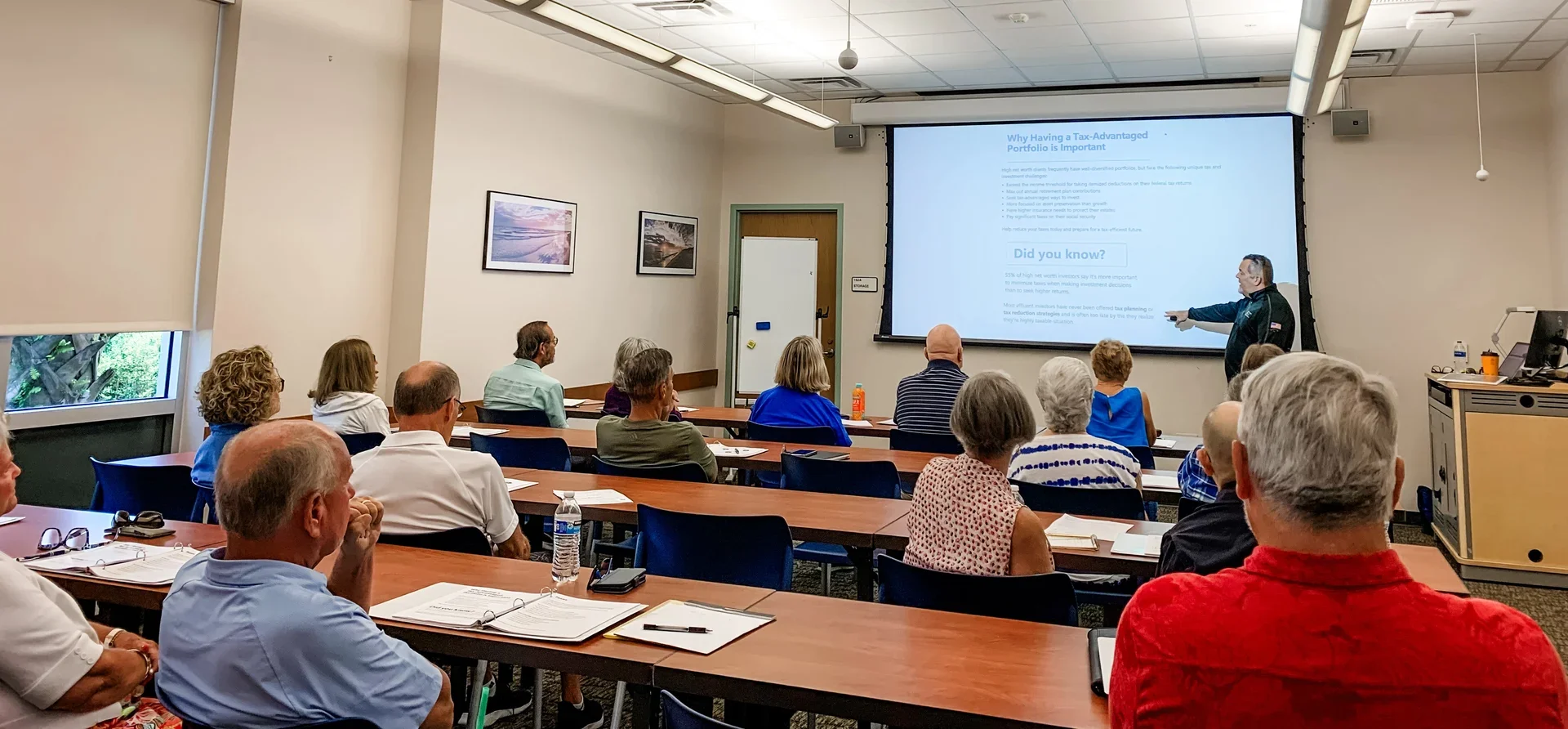 A group of people seated in a classroom, attending a presentation. The instructor is standing near a large screen displaying a slide about tax-advantaged investment portfolios.