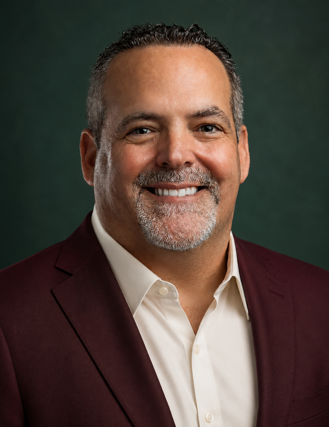 A smiling man with salt-and-pepper hair and a beard, wearing a burgundy suit jacket and a white dress shirt, against a dark green background.