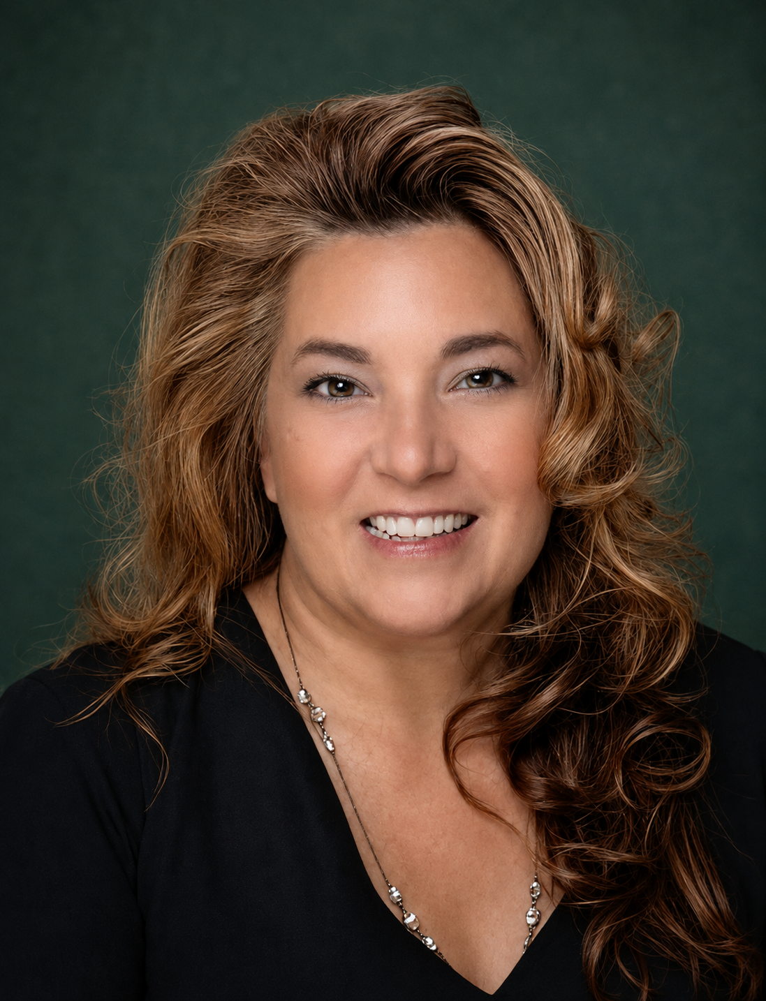 Headshot of a smiling woman with long, wavy, light brown hair, wearing a black top and a silver necklace against a dark green background.