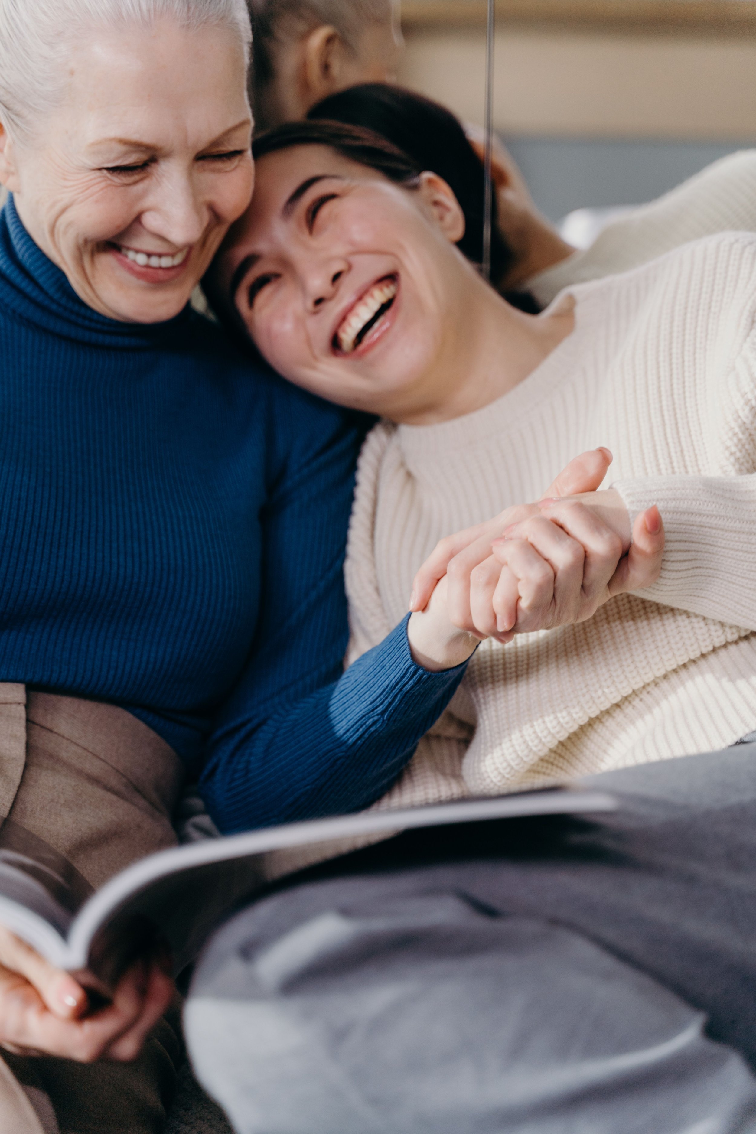 Two women smiling and leaning their heads together, holding hands, sitting closely on a couch, with a notebook in the foreground.