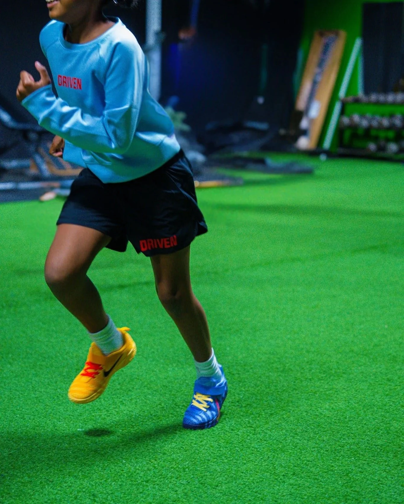 Close-up of a person running on a green indoor turf, wearing yellow and blue running shoes, black shorts with red text, white socks, and a light blue shirt with red text. The background is dark with some equipment and green walls.