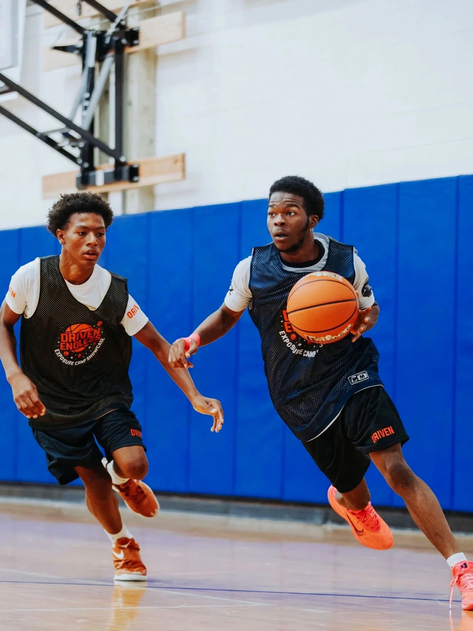 Two young male basketball players dribbling on an indoor court, wearing black jerseys with orange details, and intense expressions.