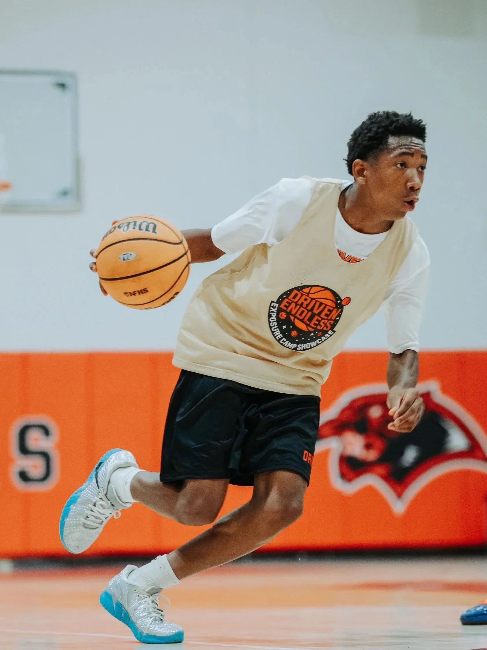 A young male basketball player in a white and yellow jersey dribbling a basketball on an indoor court with orange walls and a logo in the background.