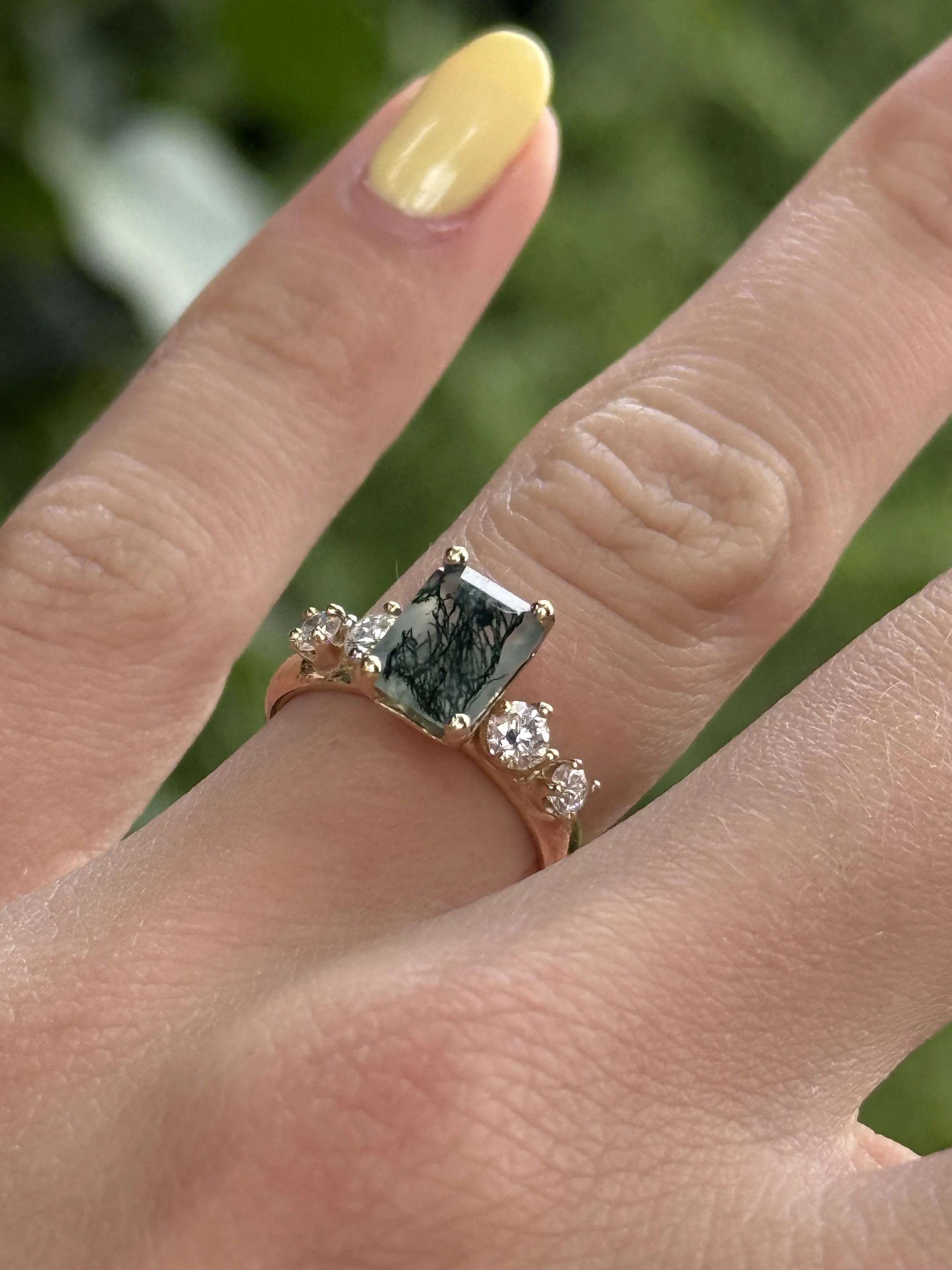 Close-up of a hand with two rings, one with a large rectangular stone with a tree-like pattern and small diamonds on the band.