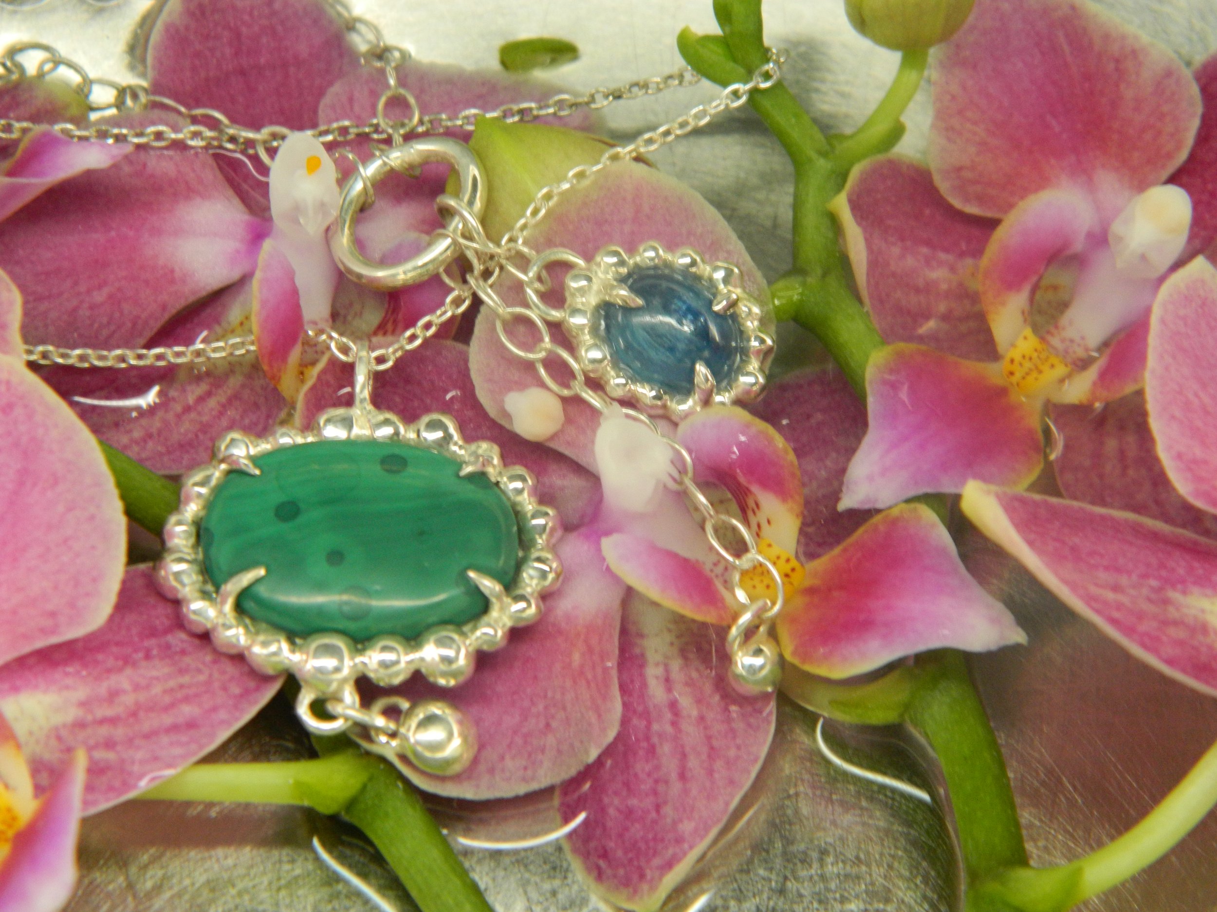 Close-up of a jewelry necklace with a blue gemstone pendant, surrounded by pink orchids with green stems.