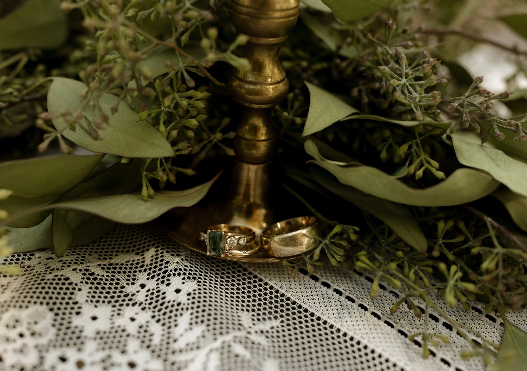 Close-up of a gold wedding band, an engagement ring with a diamond, and another gold ring on white lace fabric, surrounded by green leaves and small purple flowers.