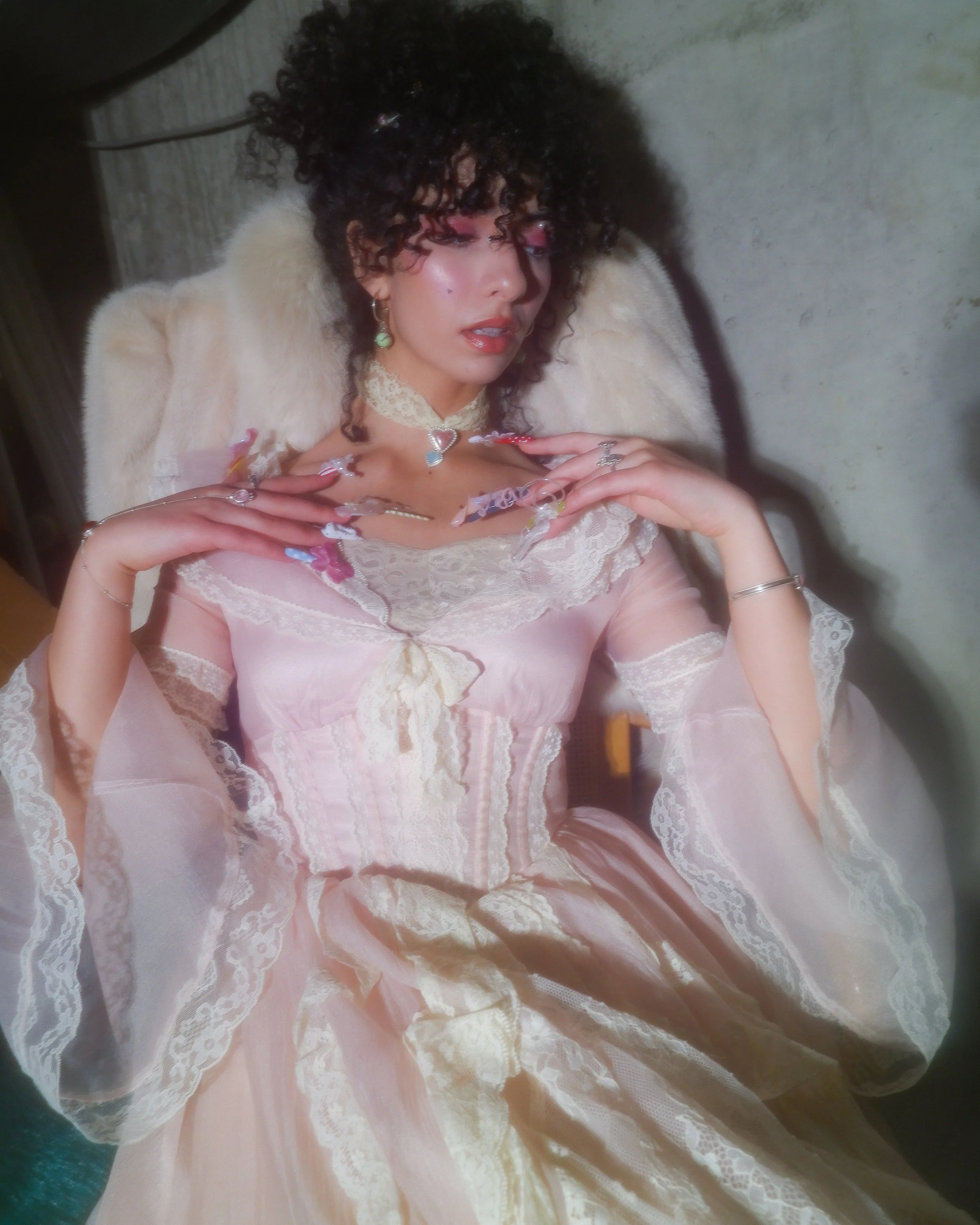 A woman with dark curly hair wearing a pink lace dress, layered necklaces, and earrings, sitting with her hands near her chest with decorated nails.