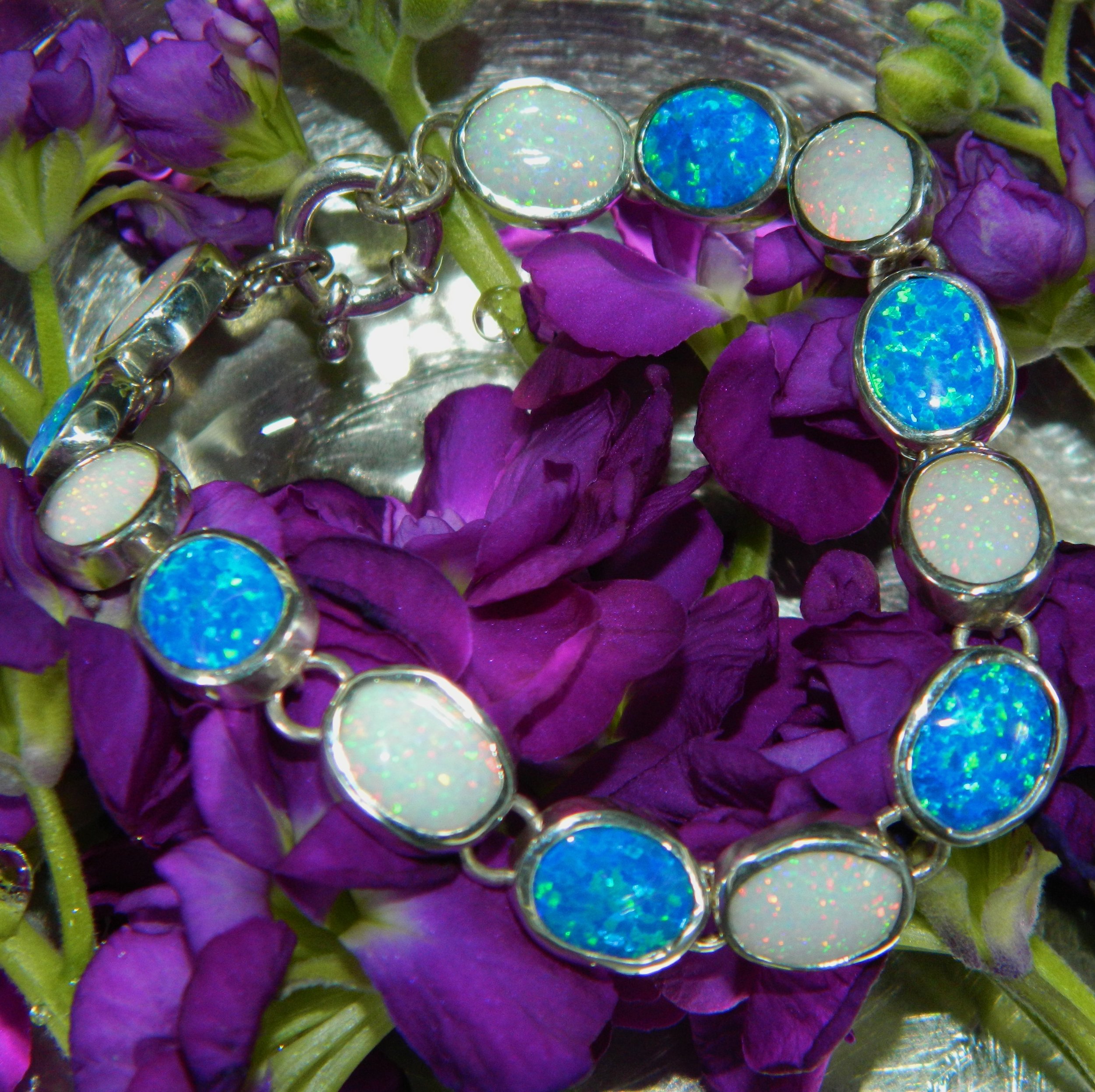 A silver bracelet with round opal and blue gemstones on a bed of purple flowers with green stems.