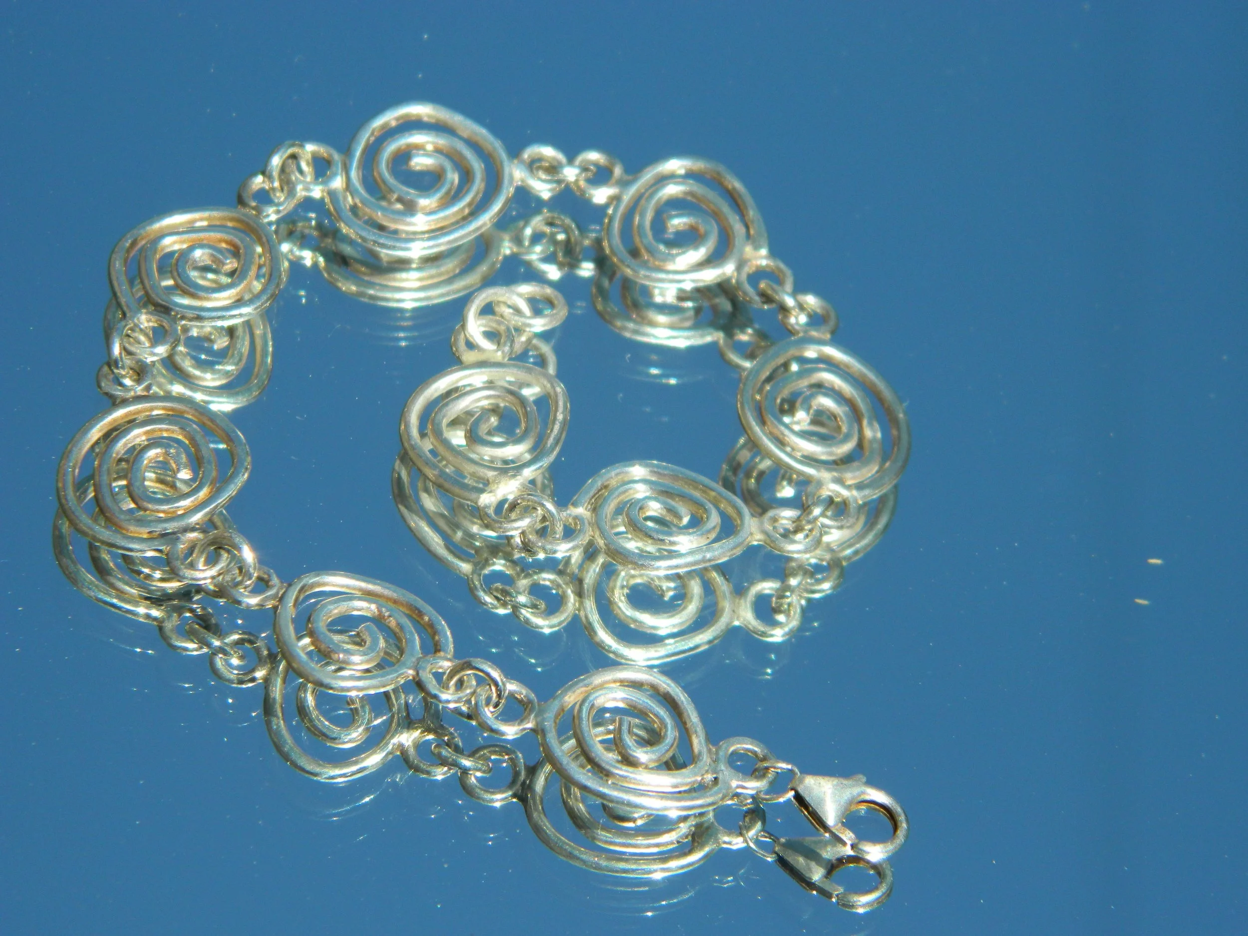 Silver chain link jewelry with spiral designs reflects on a mirror surface against a blue background.