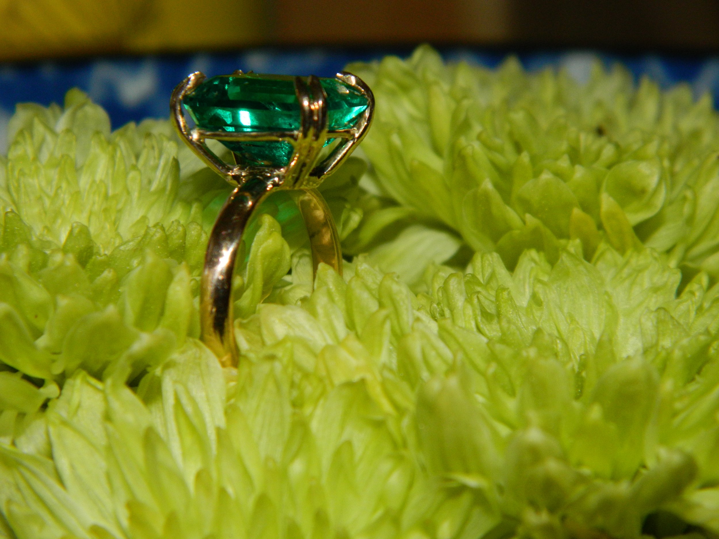 A gold ring with a large green gemstone set in a prong setting, resting on a green flower with textured petals.