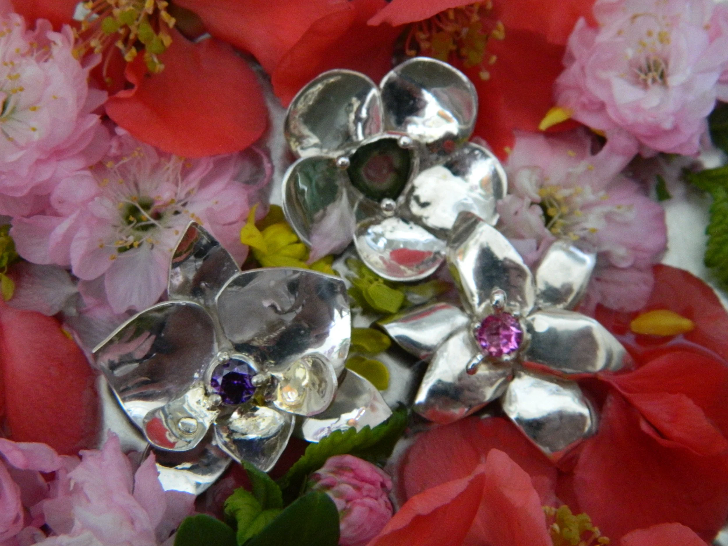 Close-up of a floral arrangement with pink and red flowers, decorated with shiny, reflective heart-shaped charms with gem-like embellishments in purple and pink, on a background of pink petals.