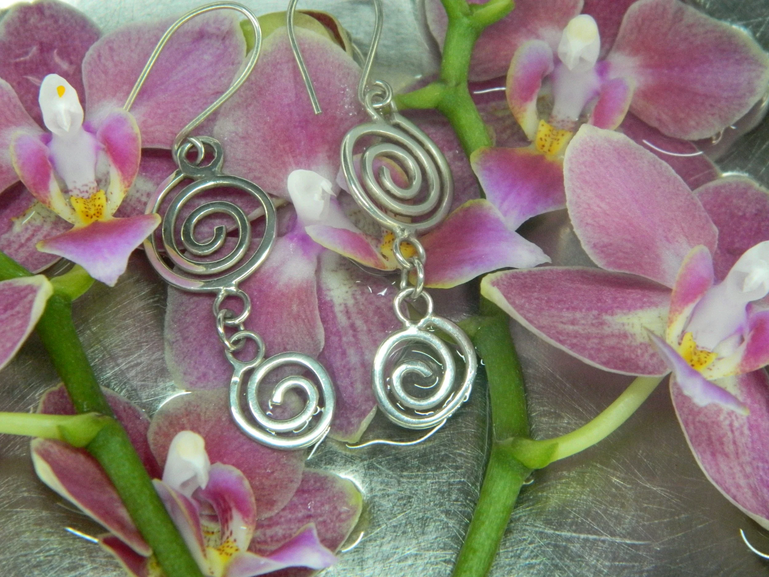 Silver spiral earrings resting on pink and white orchid flowers with green stems.