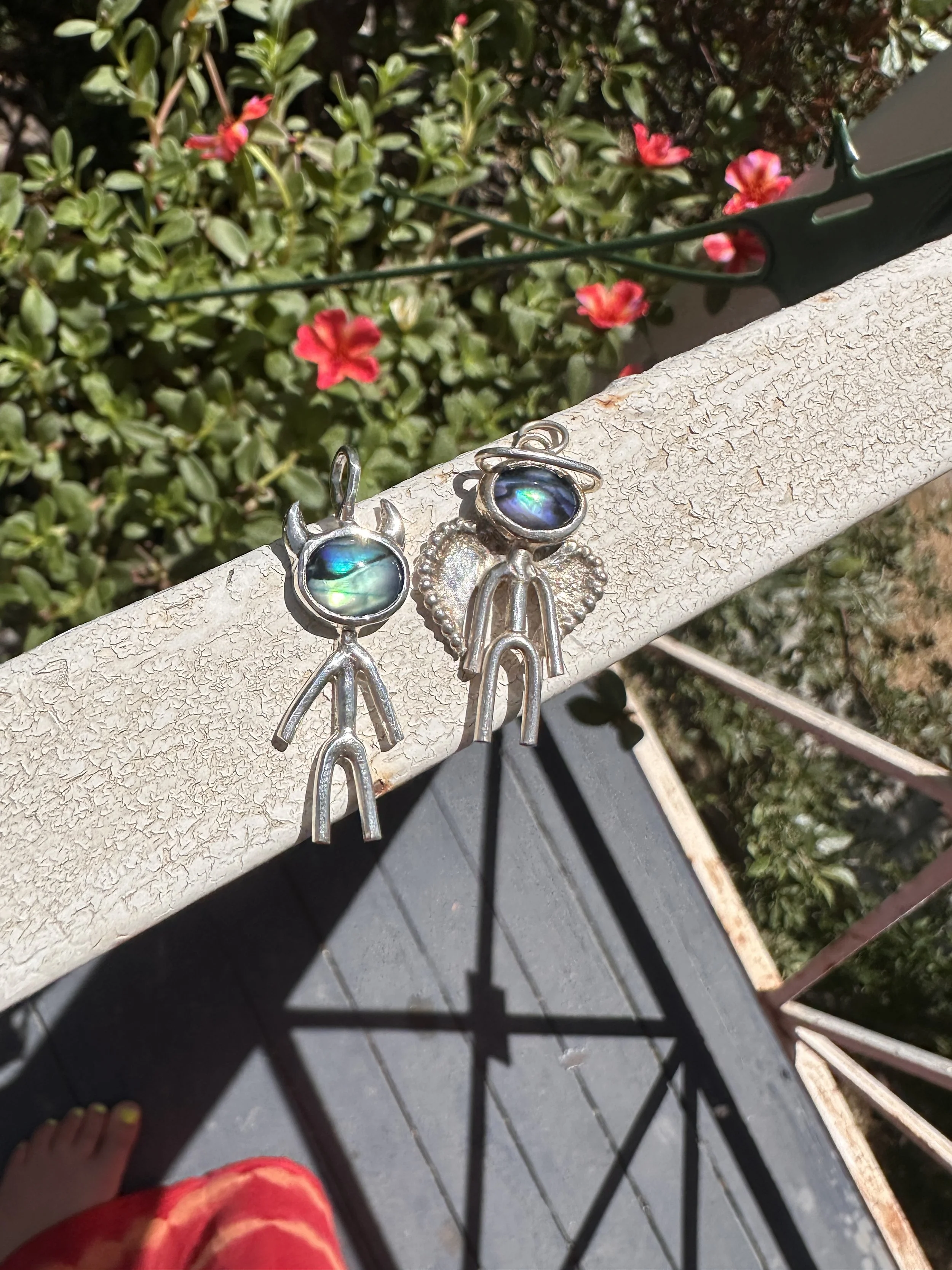 Close-up of a pair of silver angel and devil earrings resting on a textured white surface, with green bushes and red flowers in the background.