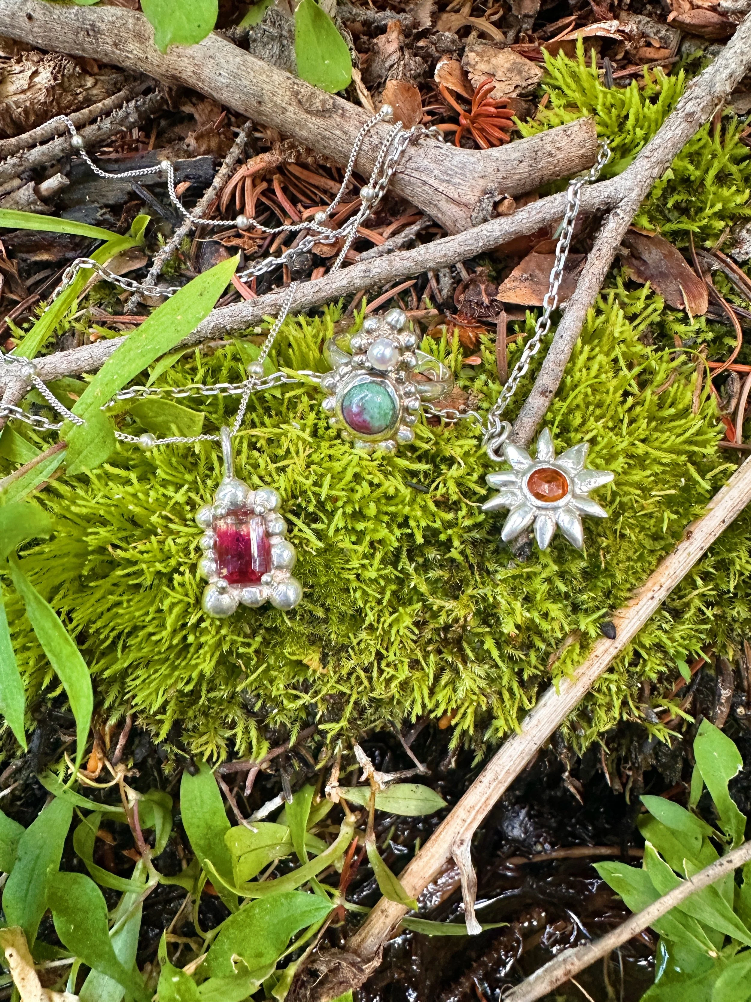 Assorted jewelry necklaces with pendants laid on moss and twigs in a natural outdoor setting.