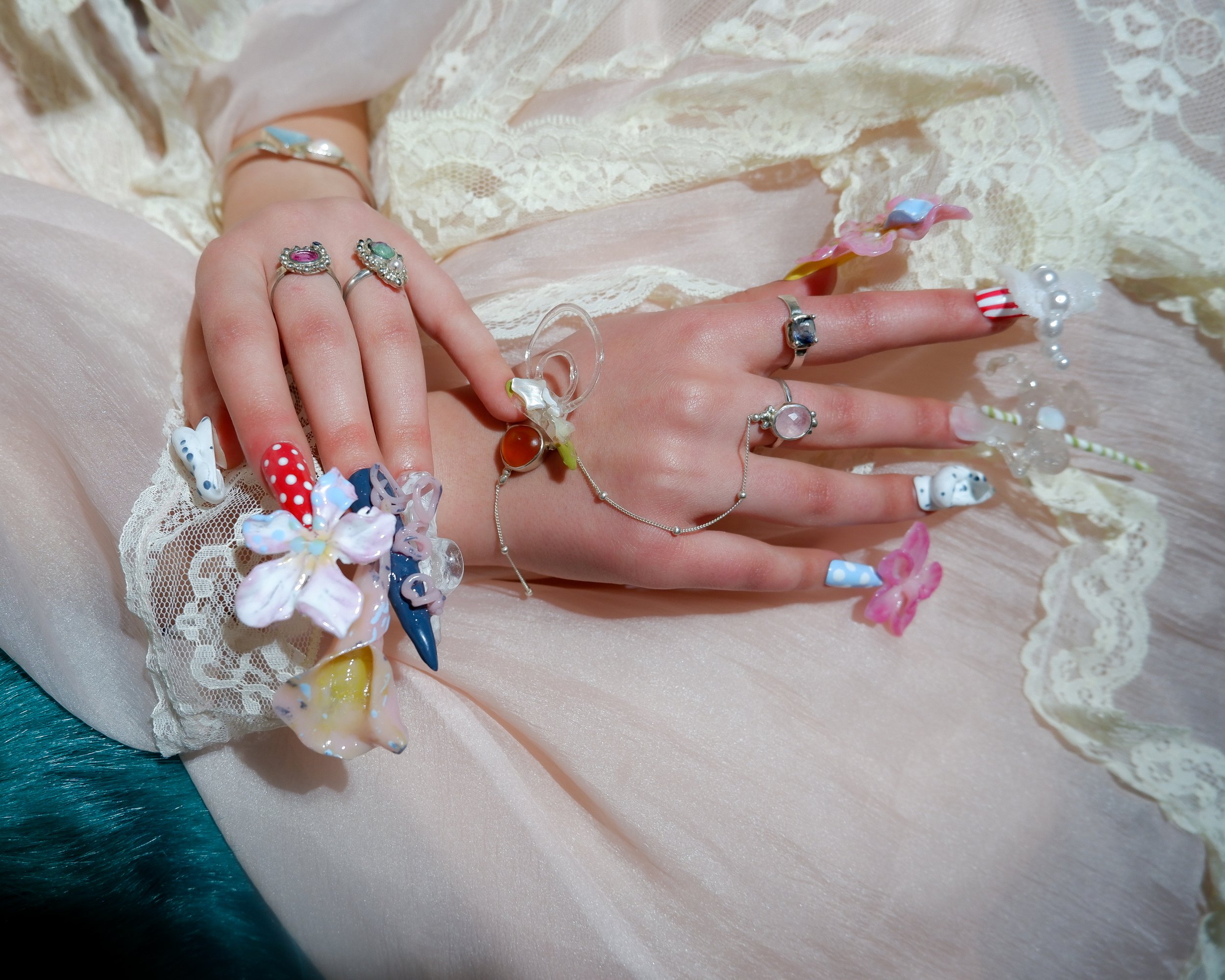 A person's hand with colorful nail art displays multiple rings and bracelets, resting on a pink lace fabric surrounded by decorative pastel-colored items and accessories.