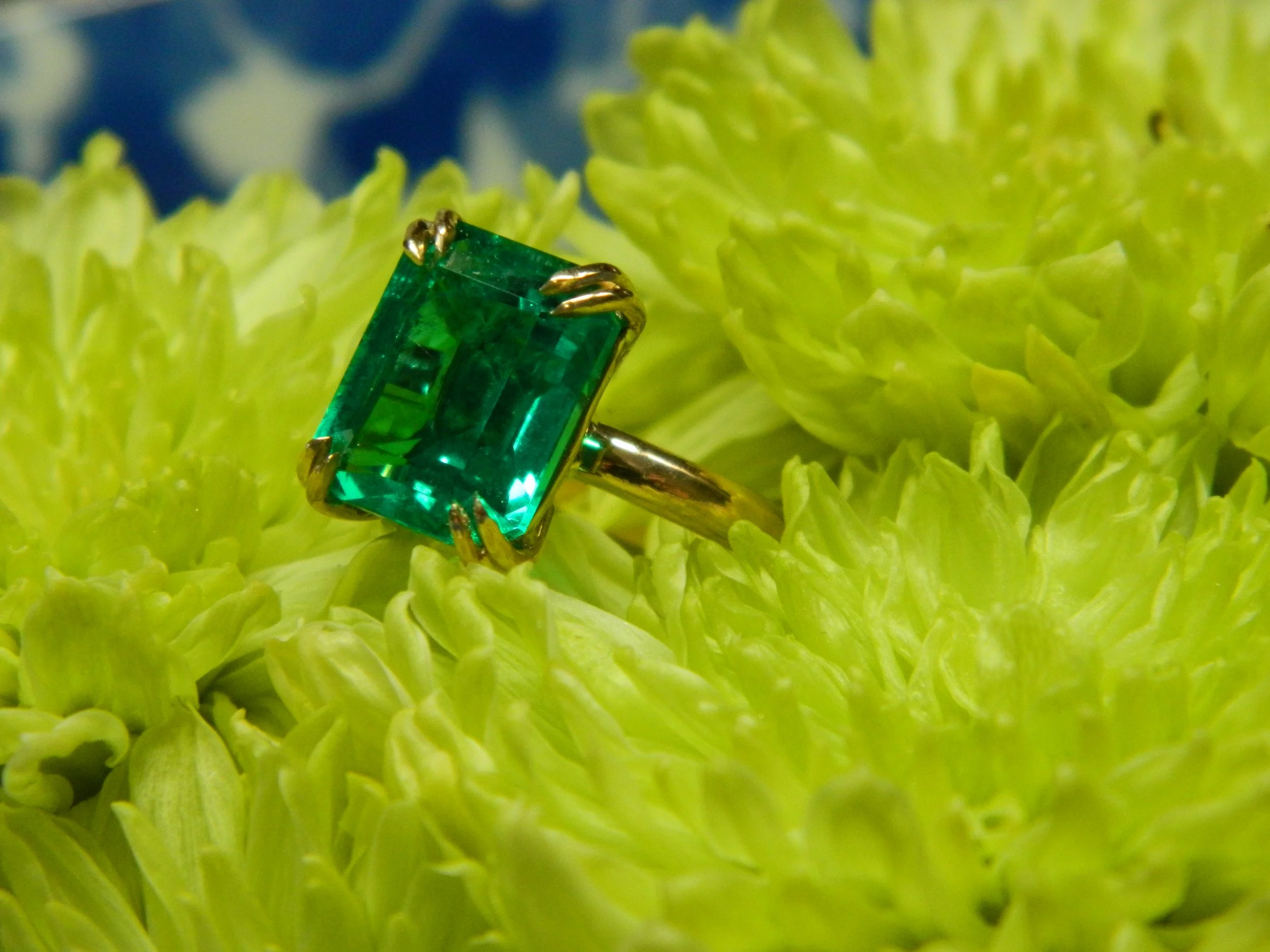 Close-up of a vibrant green ring with a large, rectangular emerald-cut gemstone set in a gold band, placed on a cluster of green plant leaves.