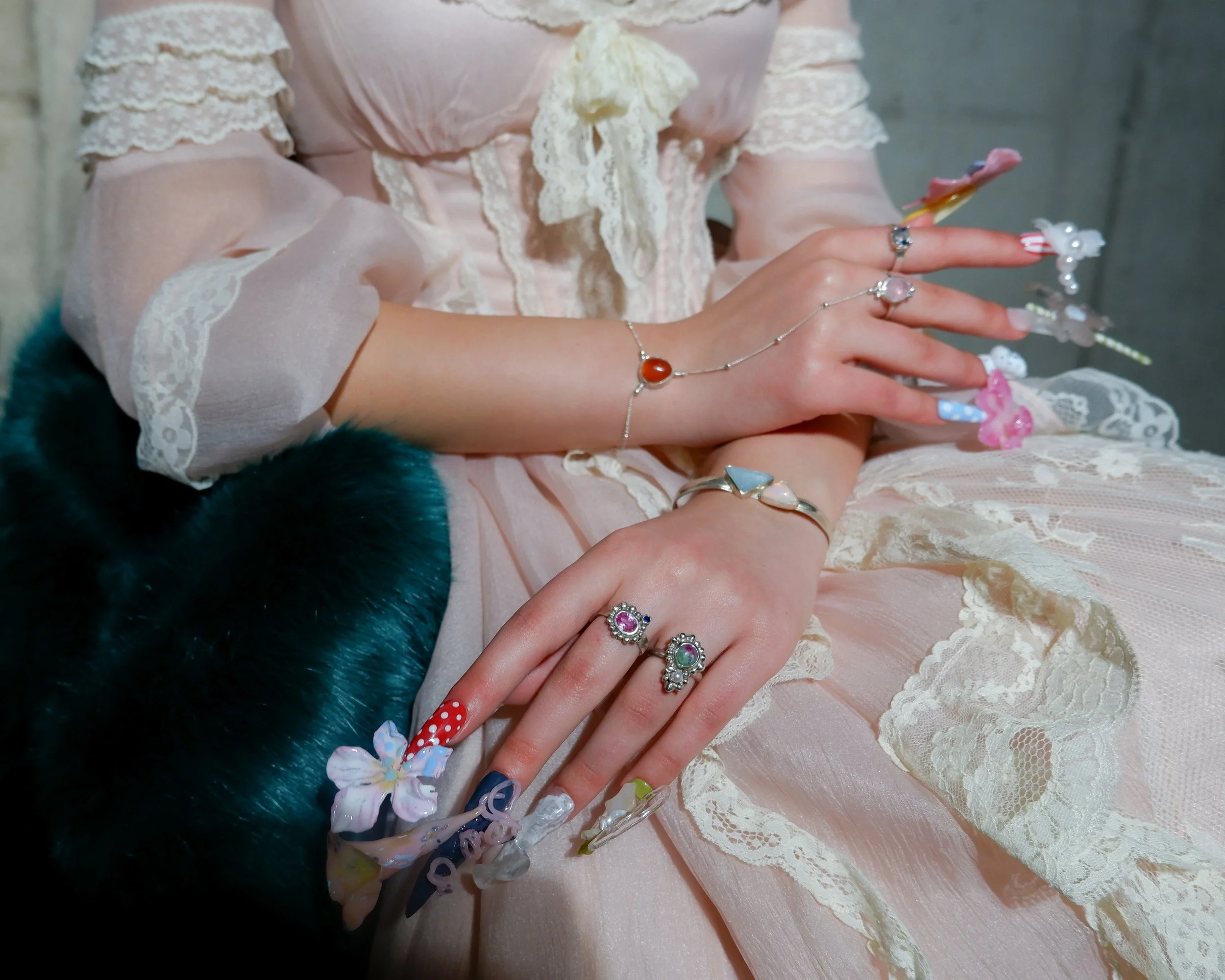 Close-up of hands wearing colorful and decorative rings and bracelets, resting on a pink lace dress with lace details on the sleeves and a bow at the collar, in front of a gray textured wall.