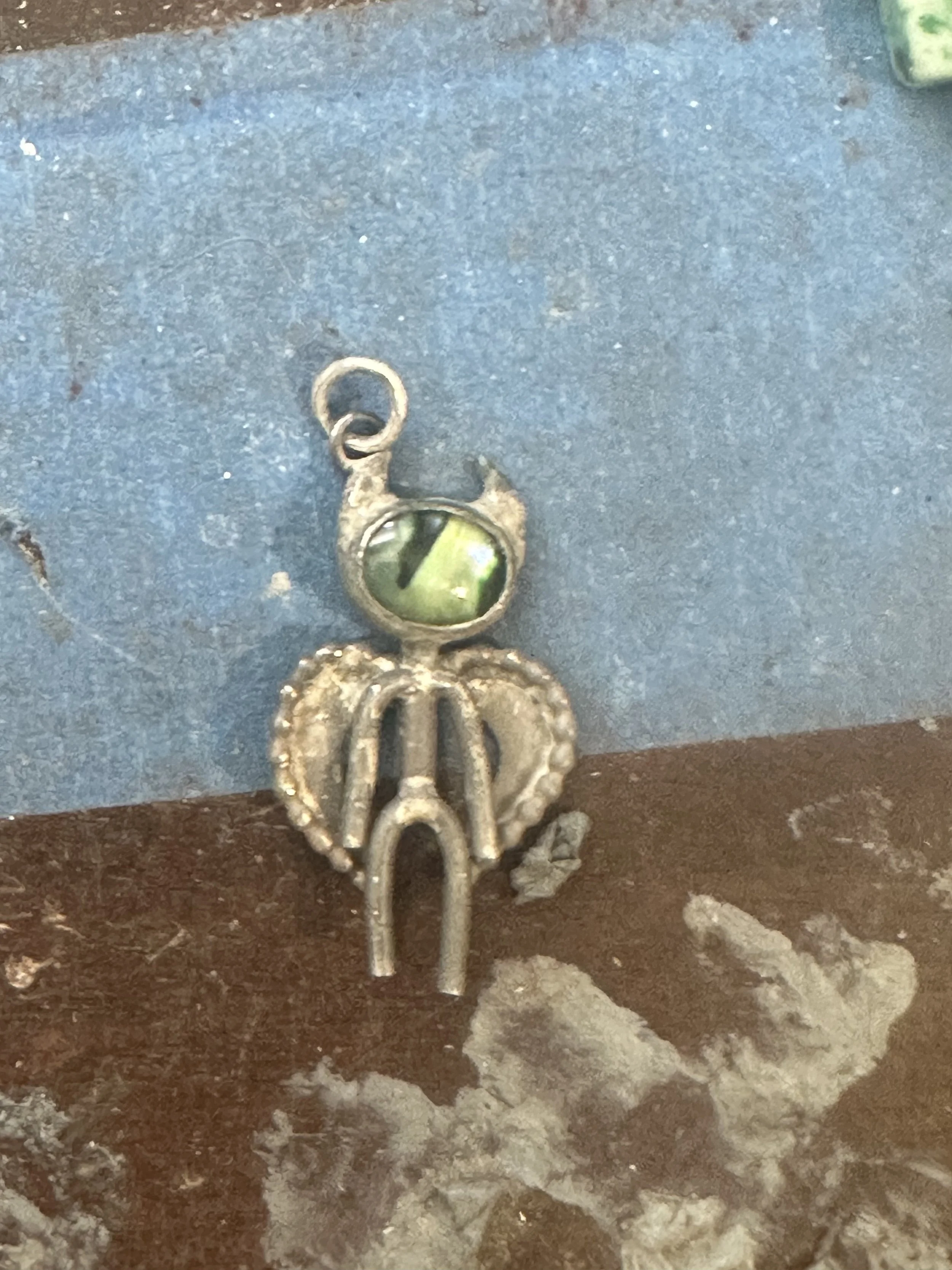 A small metal key with a decorative bow and a round head featuring a yellow-green reflective surface, resting on a textured surface along with some dried plant material and dirt.