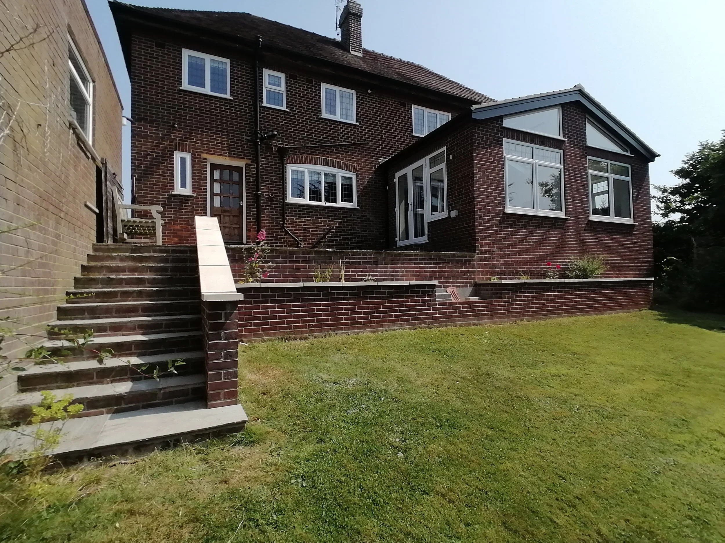 A red brick house with multiple windows, a chimney, a small staircase leading to a door, and a backyard with grass and a flower bed.