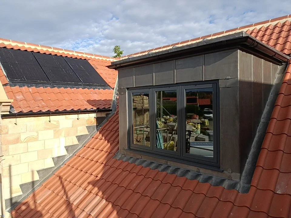 Roof with red tiles and a dormer window with black framing, reflecting outdoor furniture and plants, under a partly cloudy sky.