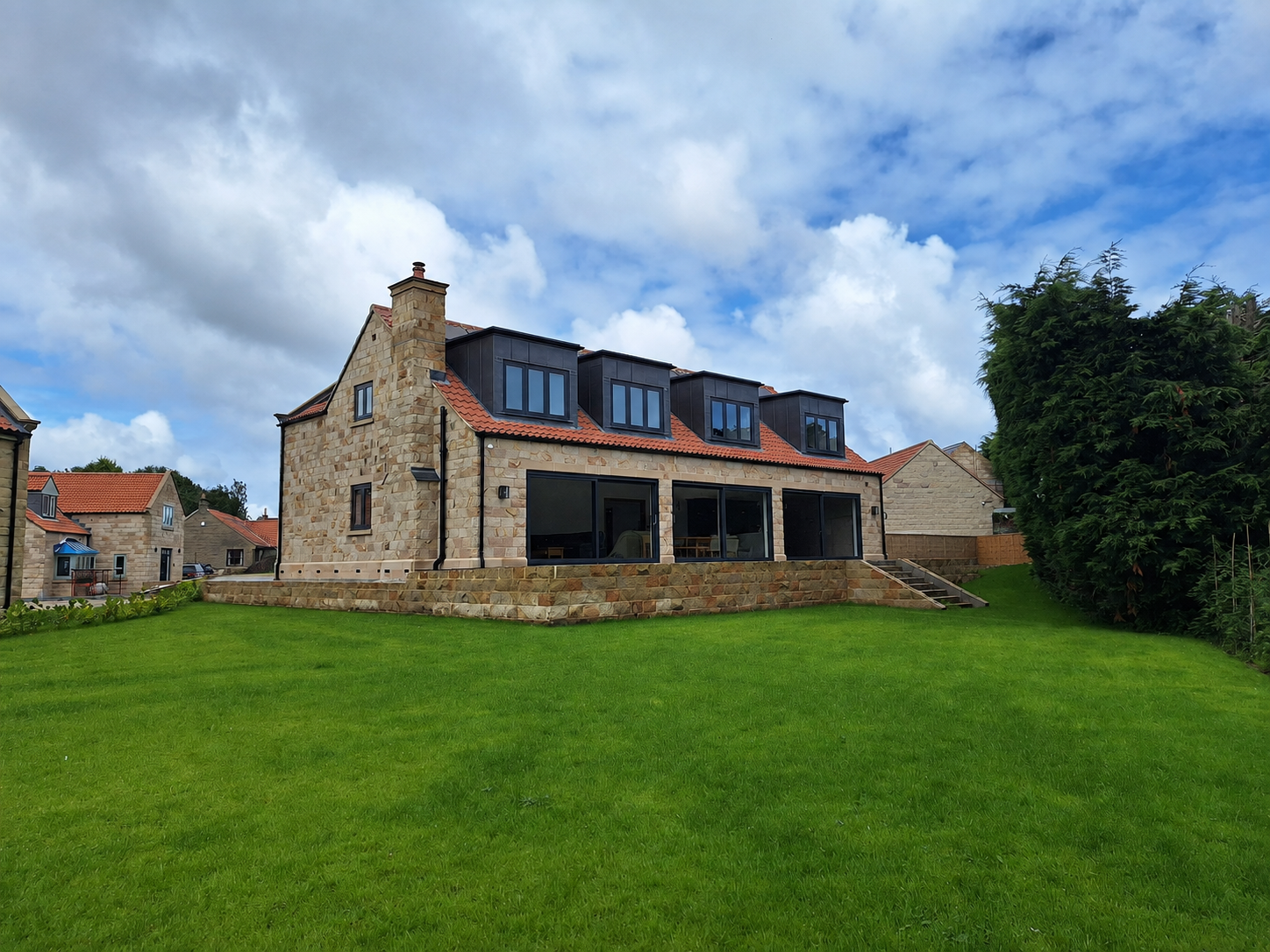 A modern house with stone walls, large black windows, and a red-tiled roof, set on a grassy lawn under a partly cloudy sky.