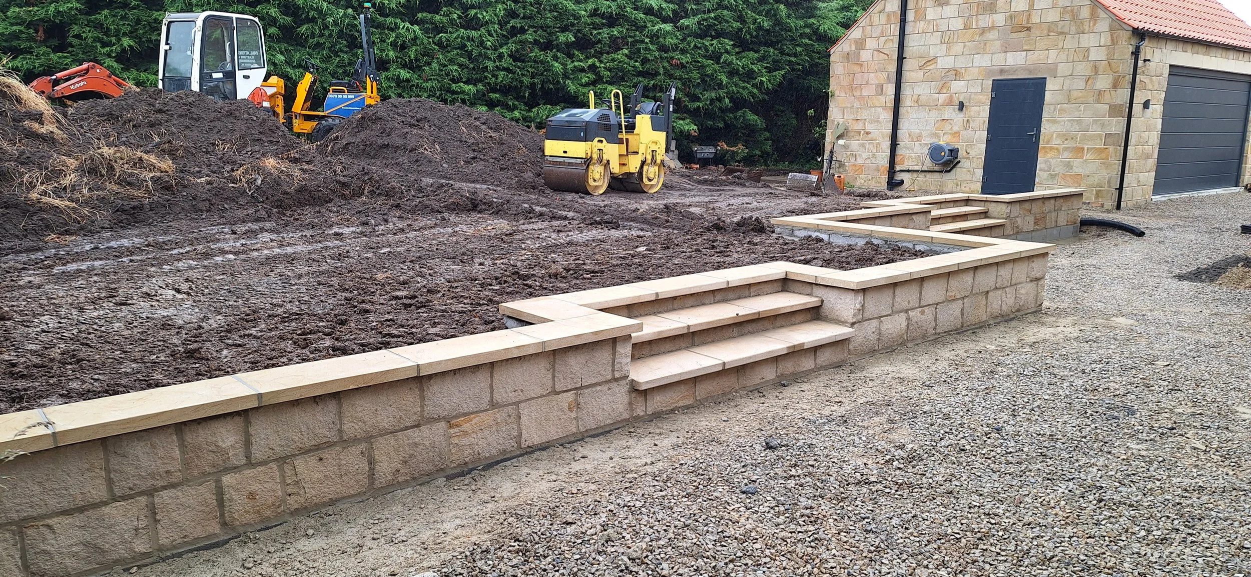 A construction site with a stone retaining wall and steps being built, dirt and construction equipment in the background, and a stone building with a garage door on the right.