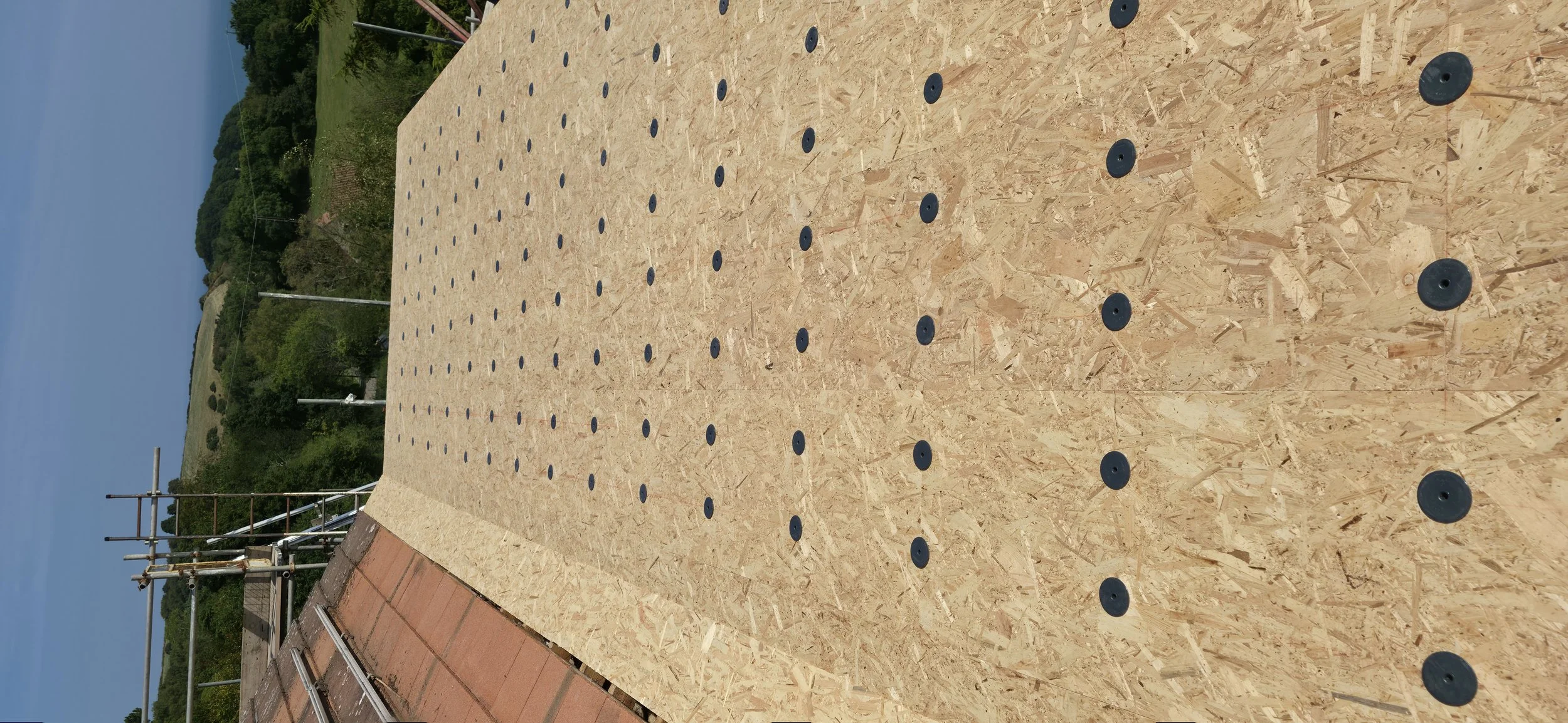 A construction site with plywood flooring and black rubber pads arranged in a grid pattern, surrounded by scaffolding and greenery in the background under a blue sky.