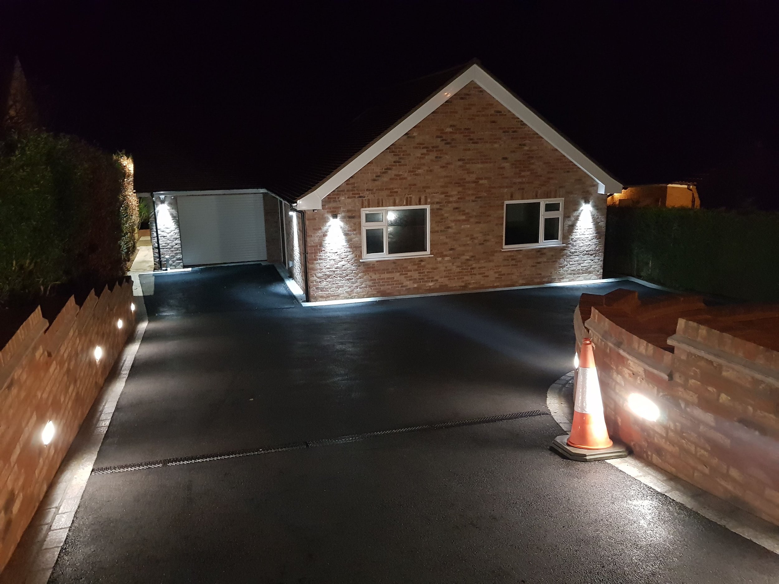 Night view of a modern brick house with exterior lights, a driveway, and an orange traffic cone in the corner.