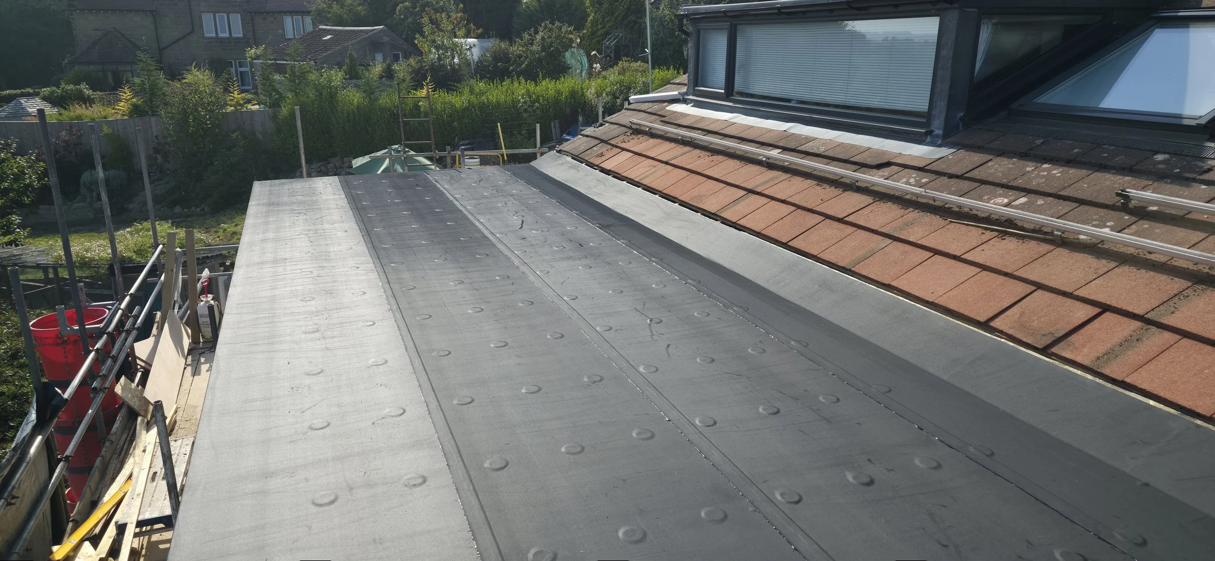 A rooftop under construction with metal and shingle roofing materials, scaffolding, and safety barriers, overlooking a residential neighborhood with trees and houses.