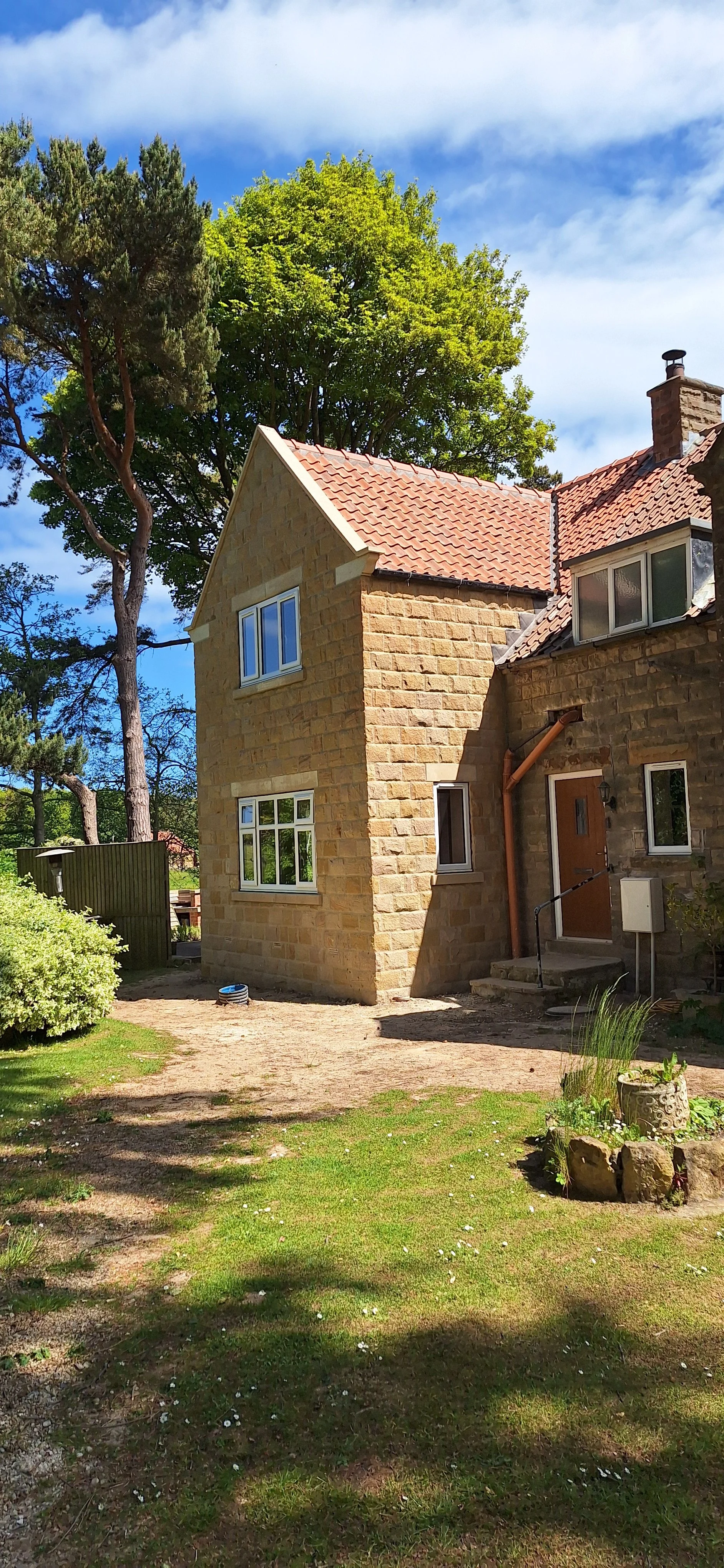 A house with a stone exterior, red tiled roof, multiple windows, and a chimney, surrounded by a garden with trees, grass, and a small flower bed on a sunny day.