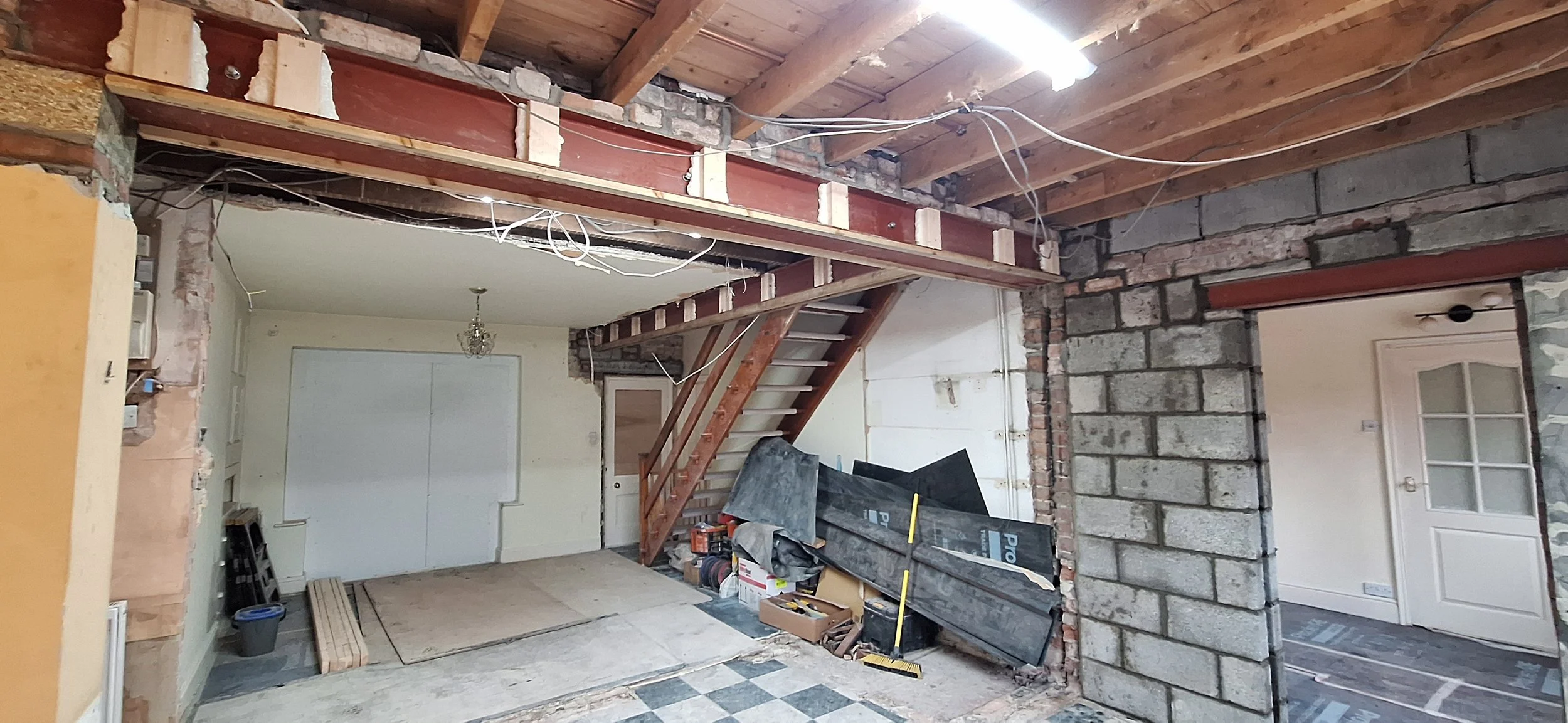 Interior of a house under renovation, showing exposed brick and wooden beams, construction materials, and a staircase leading to an upper level.