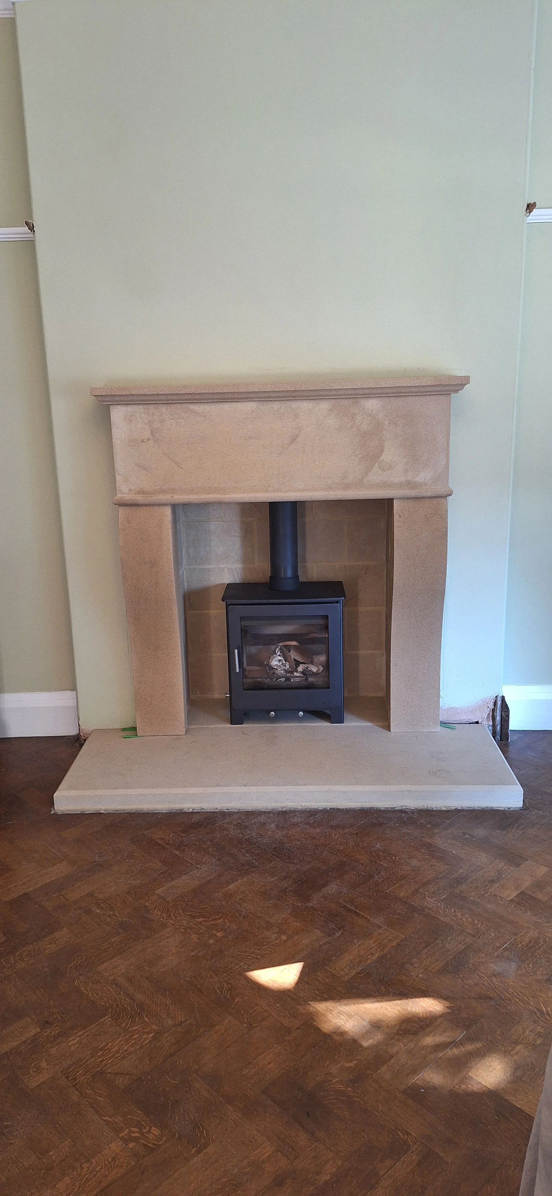 A beige fireplace with a black stove inside, installed against a light green wall with a white baseboard, on a wooden floor in a room.