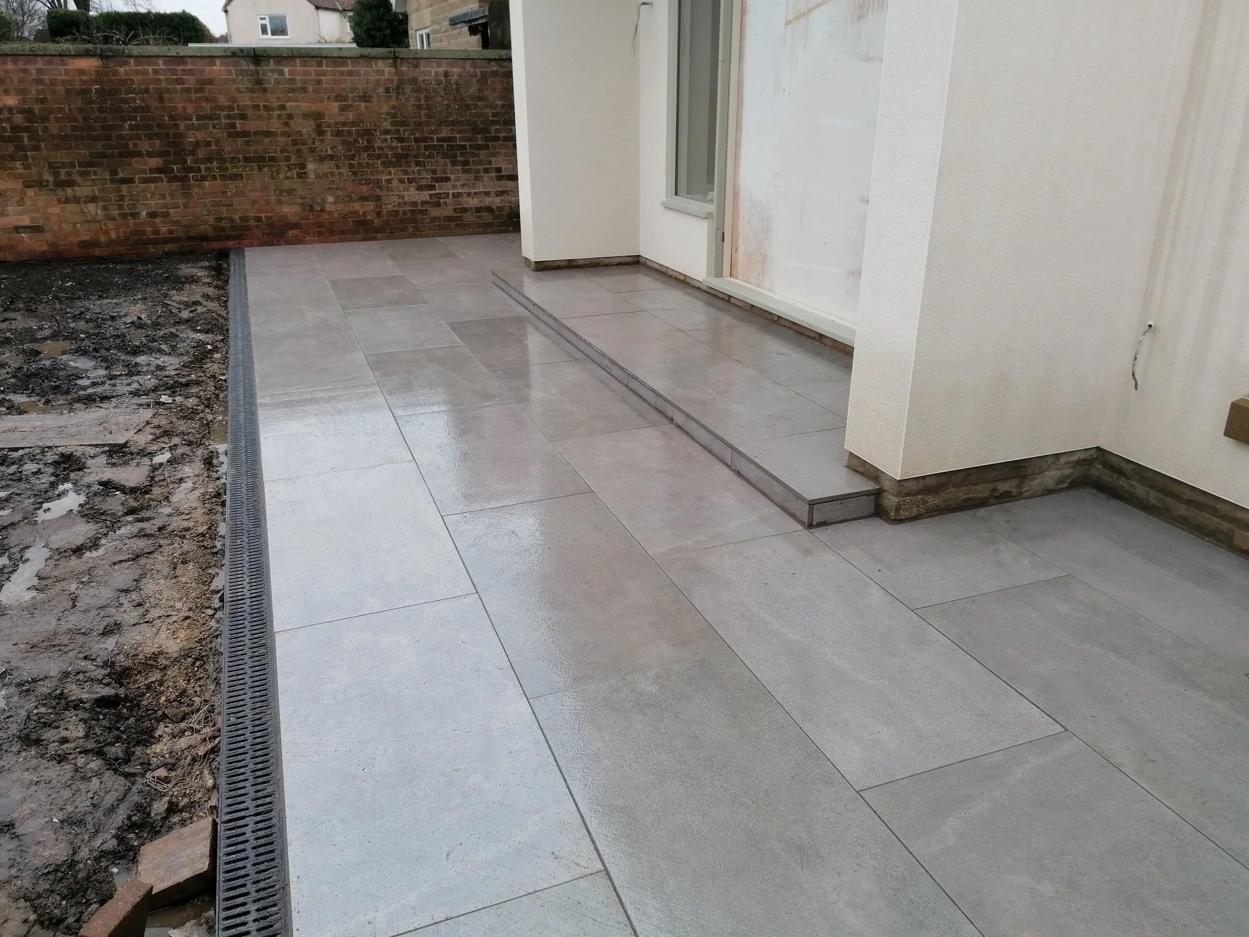A newly installed tiled patio or porch area adjacent to a white building with a door and window. The tiles are large and light in color, with a slight reflective sheen. There is a brick wall in the background and some construction or renovation work appears to be ongoing, as dirt and unfinished areas are visible.