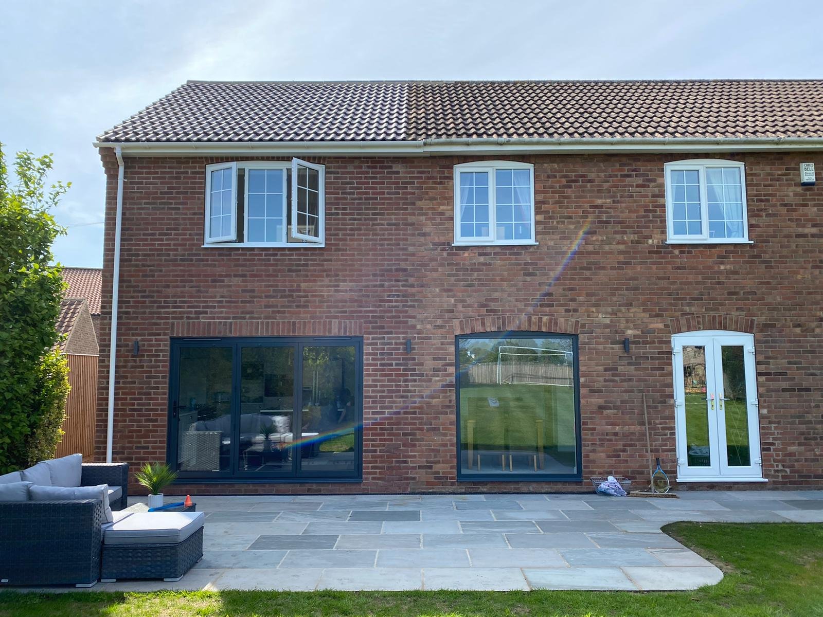 Back view of a red brick house with large glass sliding doors and two white-framed windows on the top floor, set against a blue sky with sunlight creating a rainbow lens flare. Outdoor furniture and a grassy lawn are visible in the foreground.