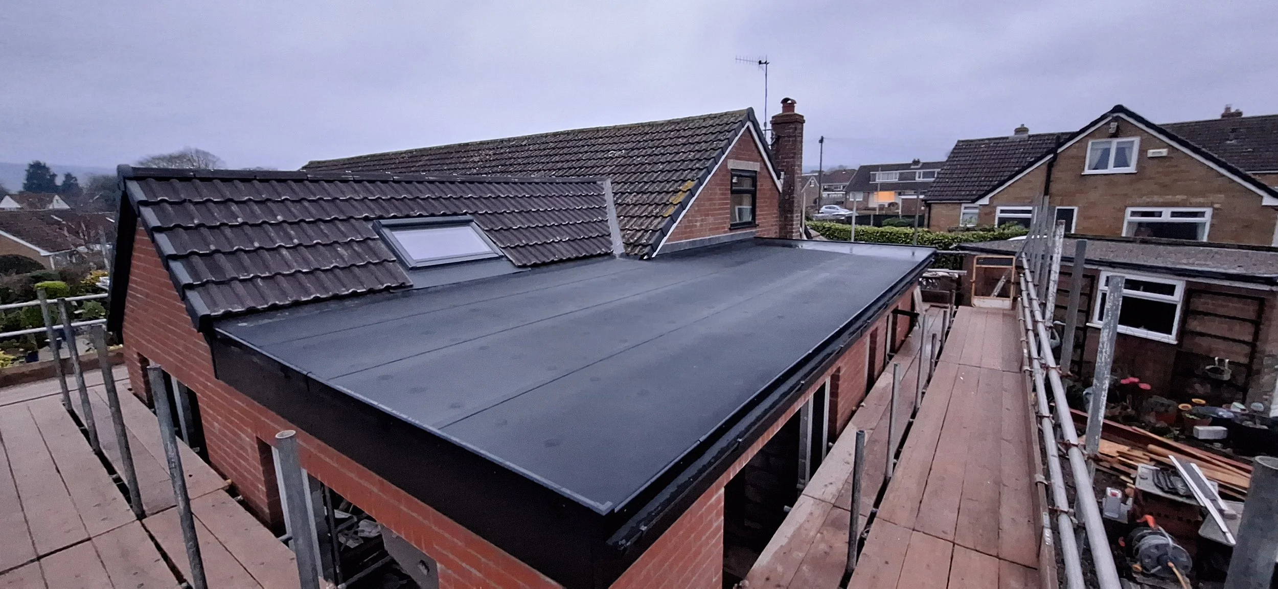A rooftop under construction with a flat black surface and a sloped tiled roof. Scaffolding surrounds the building, and neighboring houses are visible in the background on a cloudy day.