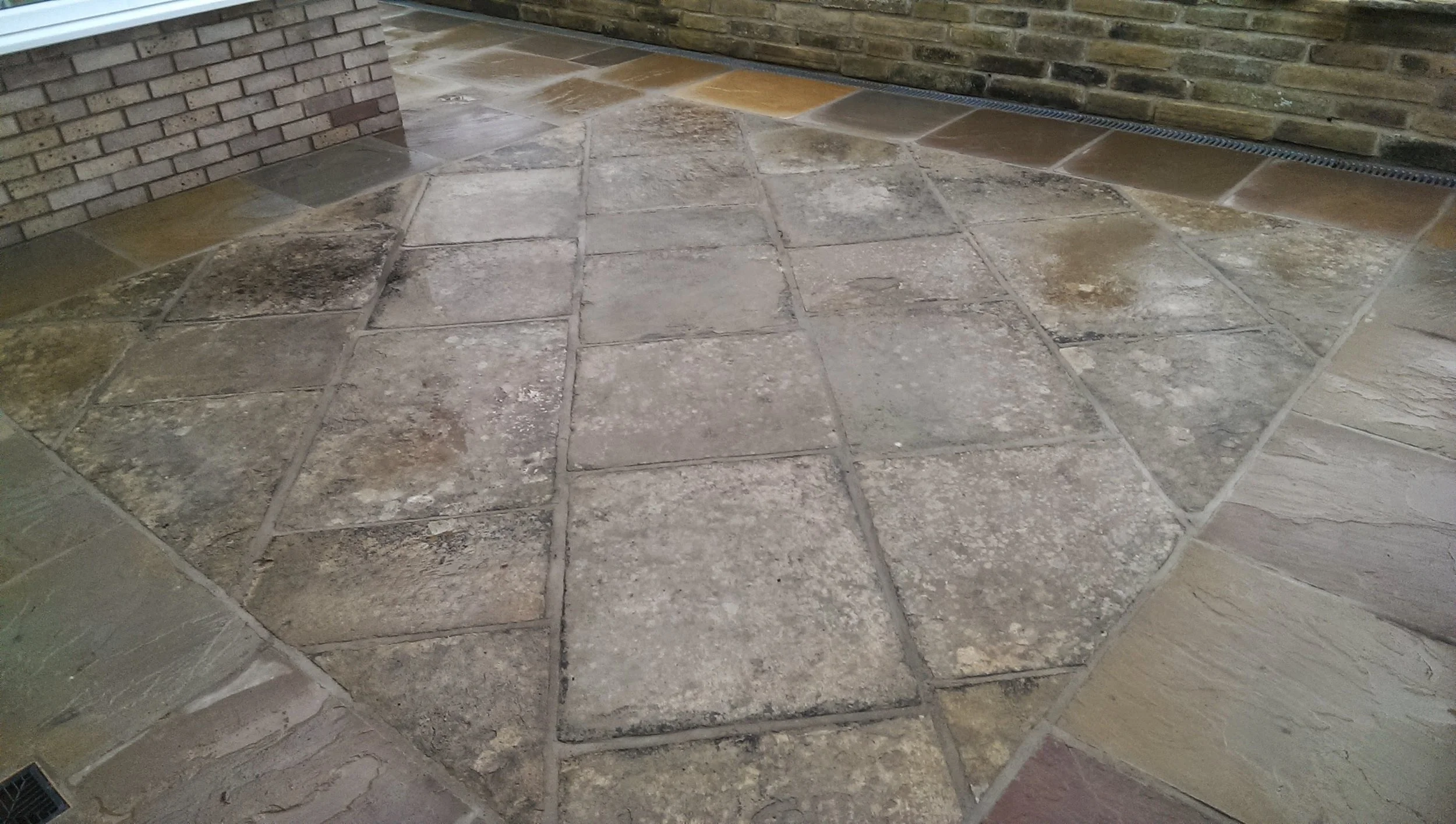 Patterned stone tile flooring outside near a brick wall and a wooden wall, with an outdoor drain grate visible.