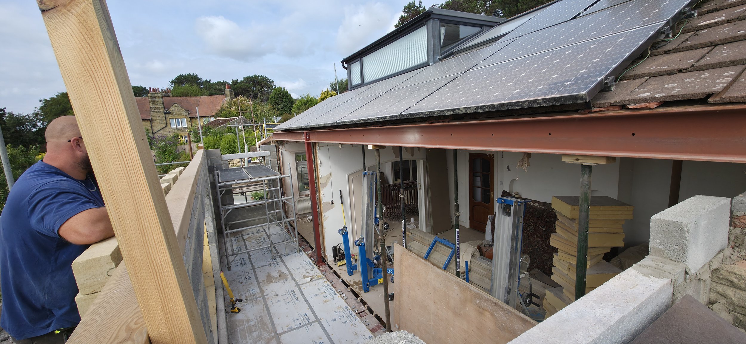 A construction worker or homeowner in a blue shirt is working on a house roof with solar panels and scaffolding, surrounded by construction materials, with a residential neighborhood and cloudy sky in the background.