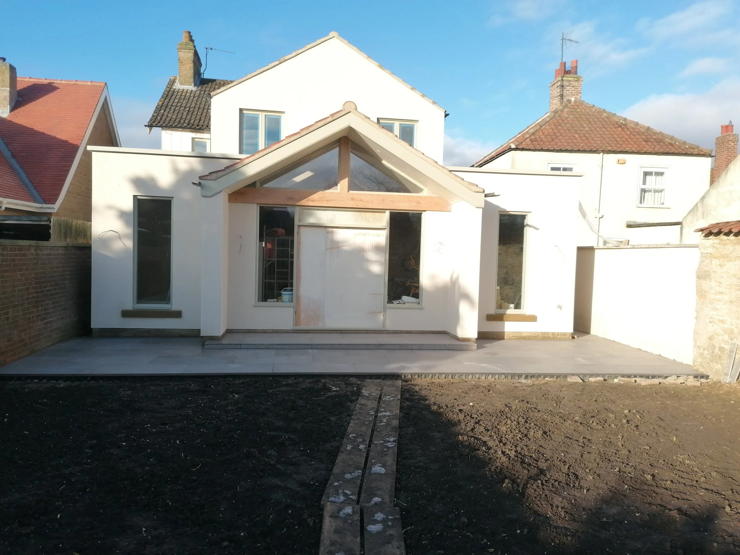 A modern house under construction with white exterior walls and large windows. The front shows a concrete patio area, and the backyard has freshly turned soil. The house features a gabled roof section with exposed wooden beams. There are neighboring houses with traditional tiled roofs visible in the background, and the sky is clear with some clouds.