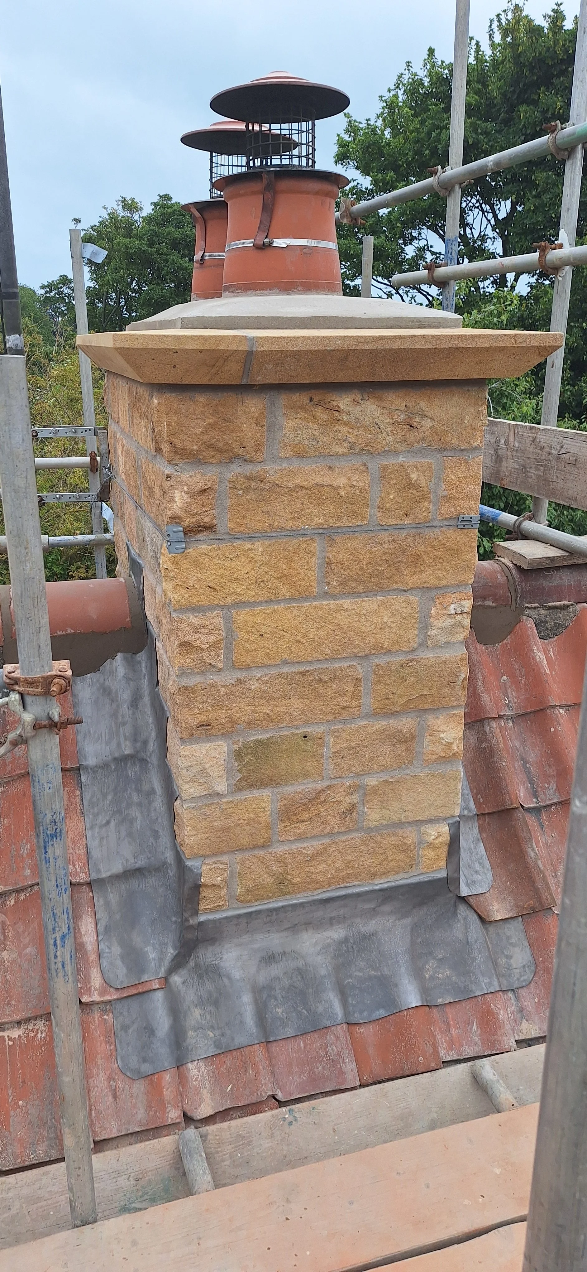 Brick chimney with chimney pots on a roof under construction, surrounded by scaffolding and green trees in the background.