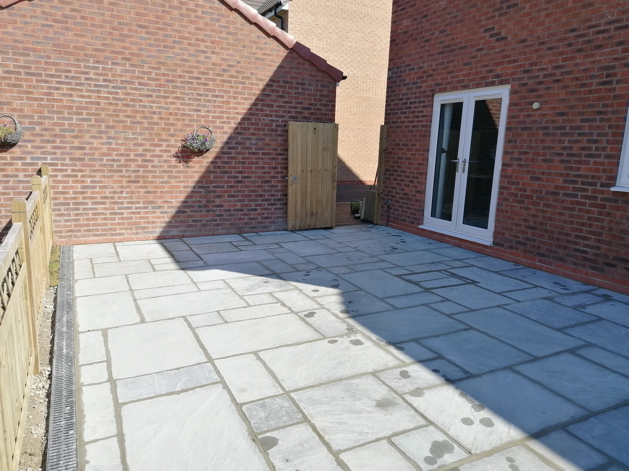 An outdoor patio area with white stone paving, a wooden fence on the left, and a red brick house with a sliding glass door on the right. There are two hanging flower baskets on the brick wall and a small wooden gate in the middle. Shadows are cast across the patio.