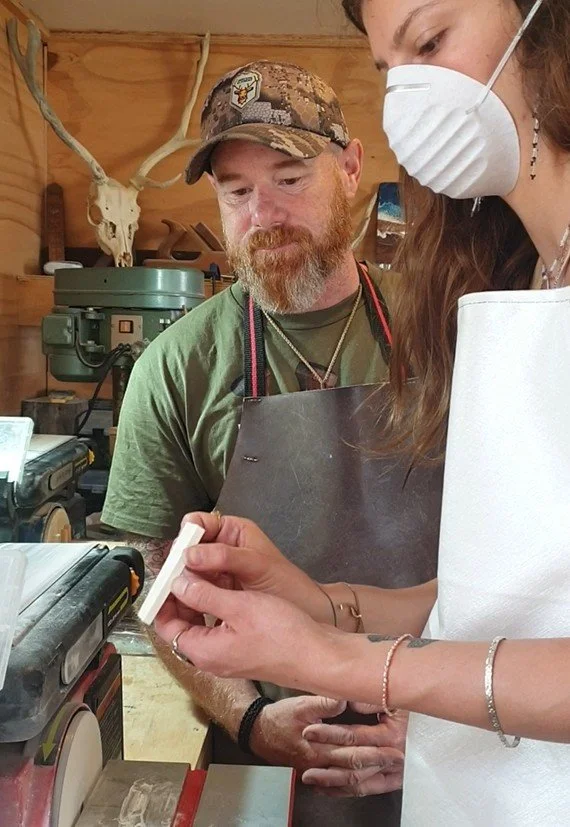 A woman wearing a white apron and face mask is showing a small object to a man with a beard and camo baseball cap in a workshop. The workshop has wood walls, a mounted animal skull with antlers, and various tools and equipment.