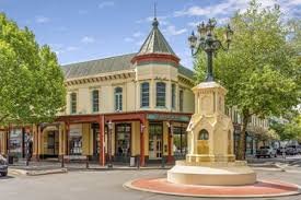A historic building with a turret, shops on the ground level, and a decorative lamp post in front, surrounded by green trees.