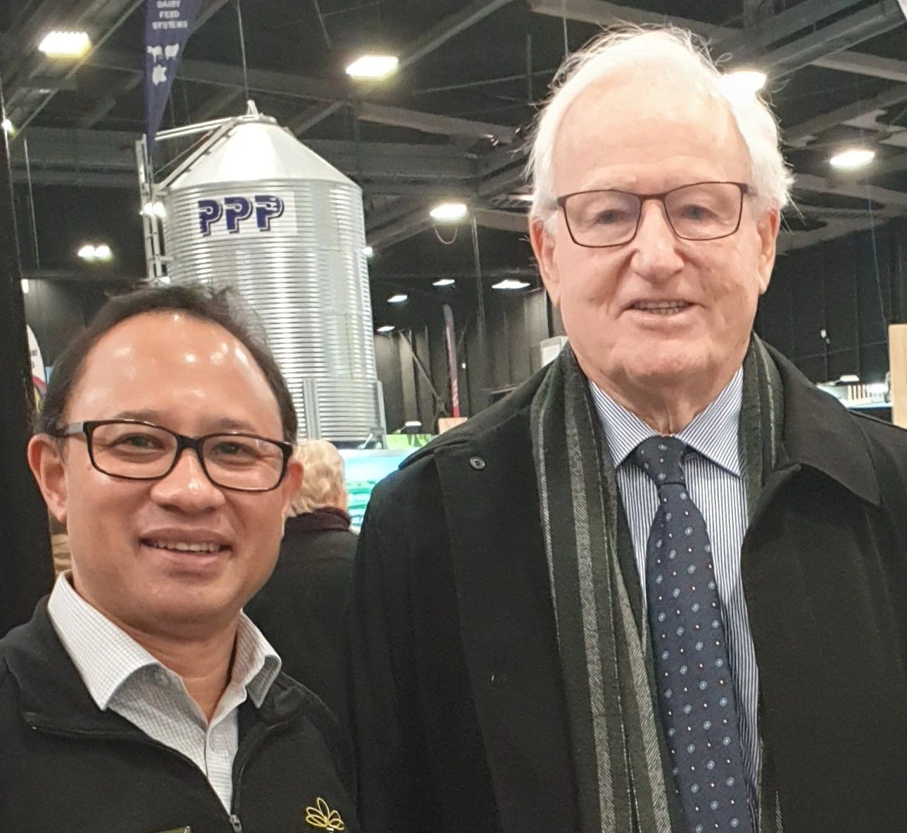 Two men standing together indoors, smiling. The man on the left has glasses and short dark hair, wearing a white shirt and black jacket with a logo. The man on the right has glasses and white hair, wearing a dark coat, striped shirt, and a polka dot tie. In the background, there is a metallic silo with the letters "PPP" on it, and a dark ceiling with lights.