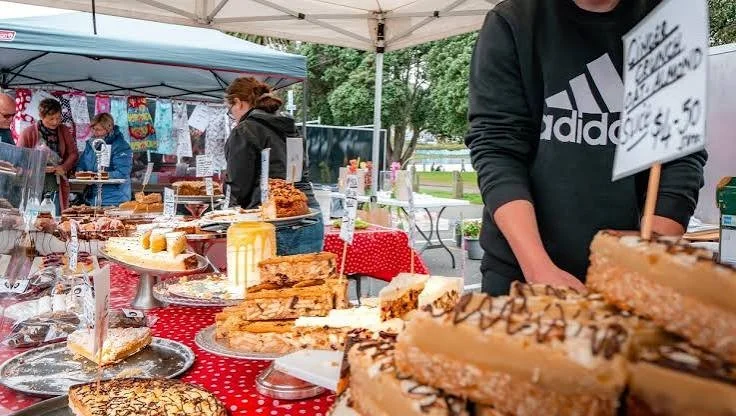 Outdoor market stall selling baked goods like cakes, cookies, and pies with people browsing and shopping.