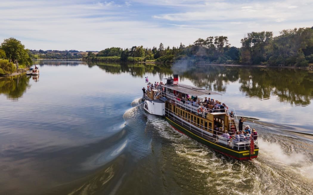 A river cruise boat sailing on a calm river with trees on both sides and a partly cloudy sky.