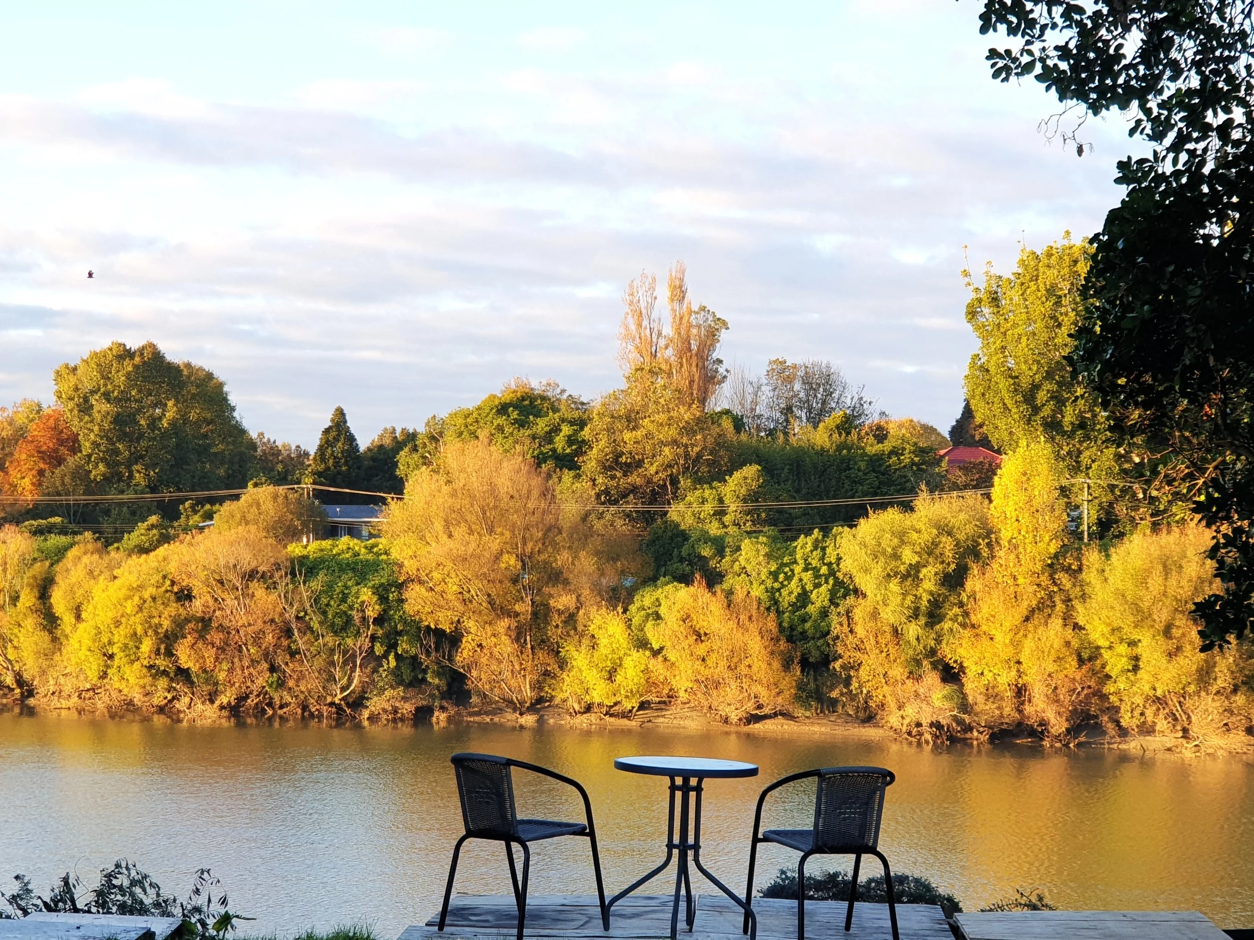 A lakeside view with two outdoor chairs and a table on a deck, overlooking a calm body of water and a lush, green forest on the opposite bank, under a partly cloudy sky.